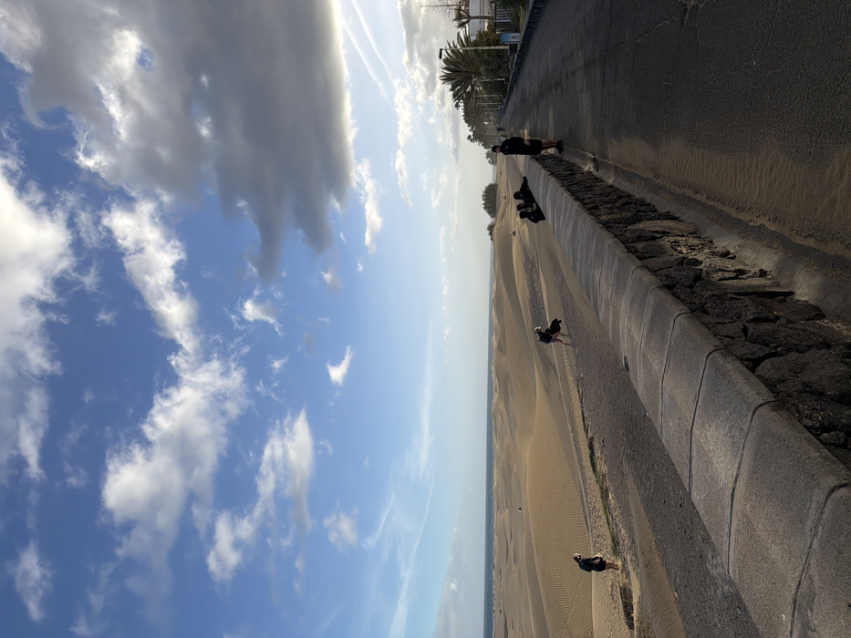 The image depicts the Maspalomas Dunes, located in San Bartolomé de Tirajana, Gran Canaria, Spain.
The scene presents an expansive landscape under a sky featuring blue patches interspersed with white cumulus clouds, transitioning to a dense mass of darker grey clouds in the upper right quadrant. High-altitude contrails are visible in the upper left. The horizon line reveals the Atlantic Ocean beyond a vast expanse of light tan sand dunes, characterized by undulating forms and visible surface ripples.
In the foreground, a low stone wall, approximately 0.75 meters high, constructed of dark, irregular stones with a grey, flat-topped cap, separates a sandy path from a dark asphalt promenade or road. The promenade extends into the distance on the right, lined by palm trees, low scrub vegetation, and streetlights. Distant white buildings are partially visible behind the vegetation.
Multiple individuals are present in the scene:
1. One person in dark attire stands on the sandy path in the left foreground, facing towards the dunes.
2. Another individual, wearing a light-colored top and dark bottoms, walks across the dunes from left to right, carrying a dark object.
3. A group of three individuals, all dressed in dark clothing, are seated or leaning against the stone wall on the dune side, facing the dunes in the mid-ground.
4. A fourth individual, wearing a dark jacket and beanie, stands on the asphalt promenade in the right foreground, positioned next to the wall and facing slightly towards the left.
The natural light suggests a low sun angle, casting shadows and highlighting the textures of the dunes and the individuals. No specific interactions between the individuals are discernible.