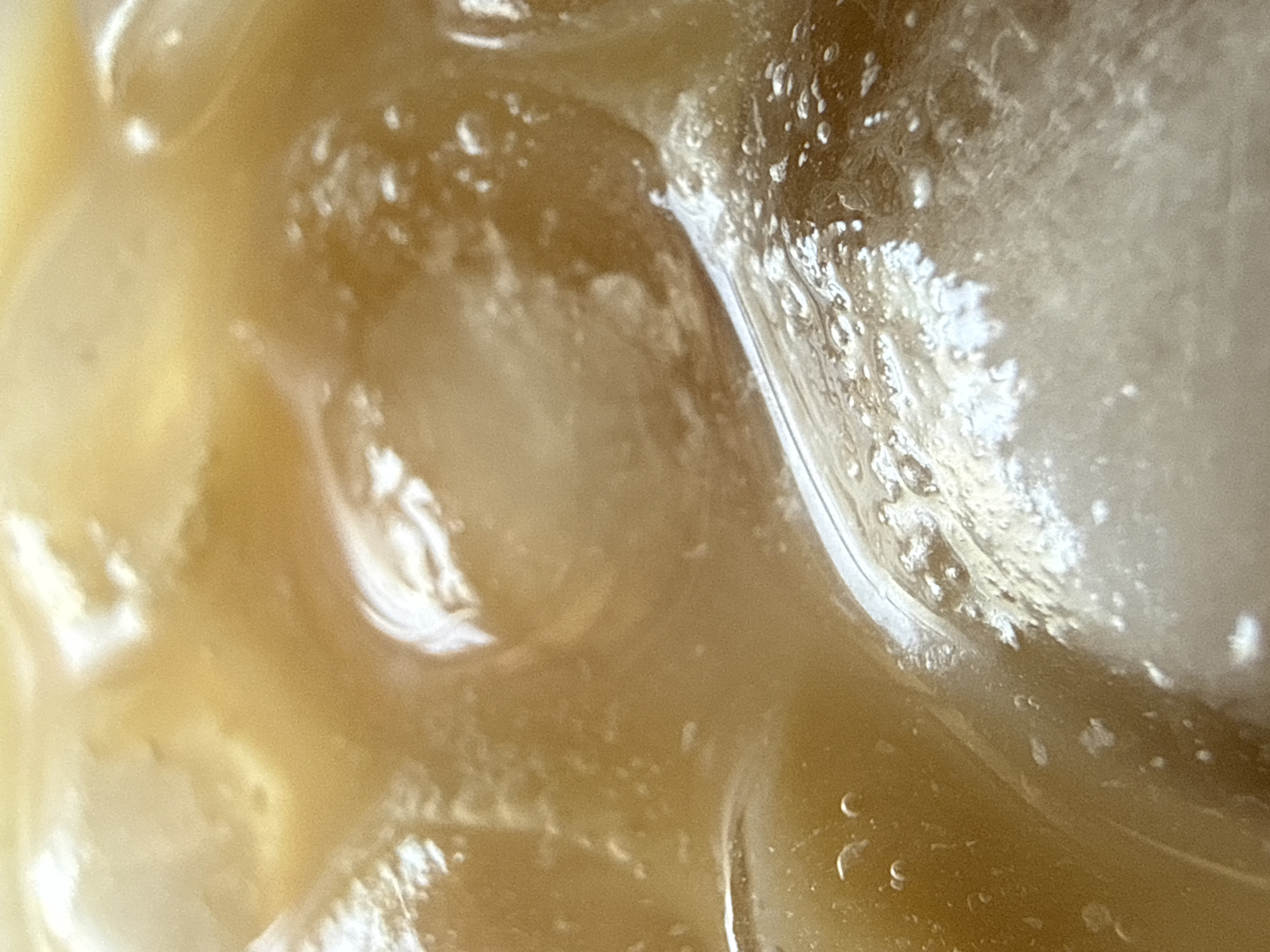 An extreme close-up, macro-level image depicts a beverage containing multiple ice cubes. The liquid is an amber to light brown hue, suggesting a beverage such as iced coffee or tea. Its surface exhibits subtle reflections and scattered small air bubbles. Numerous irregular ice cubes, appearing in various stages of melting, are submerged and partially submerged within the liquid. Their surfaces feature prominent white highlights from reflected light, indicating wetness and glossiness, alongside areas of cloudiness characteristic of ice. Small clusters of effervescent bubbles adhere to the ice surfaces and are suspended in the surrounding liquid, particularly around the points of contact and melting. The composition is tightly cropped, filling the entire frame with the liquid and ice, providing no visible edges of a container or any external background elements. No individuals or additional objects are visible within the scene. The location depicted is unidentifiable and not ascertainable from the image data.