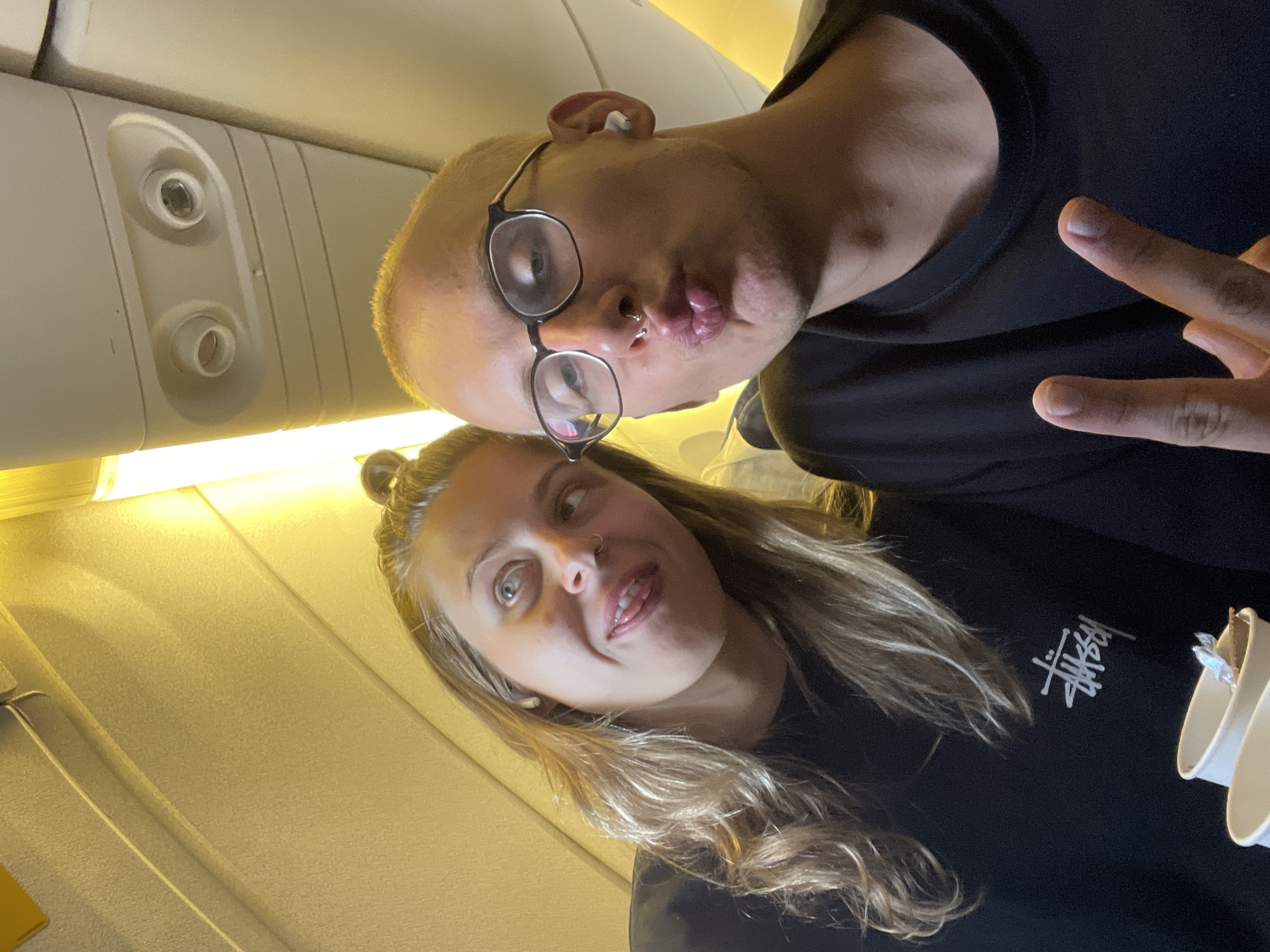 A photograph taken inside an airplane cabin features two individuals. The person in the foreground, likely male, is bald and wears glasses. They are making a puckered-lips expression while giving a peace sign, and an earbud is visible in their ear. A nose ring is also visible. Behind them, a woman with long, wavy blonde hair pulled up in a bun is smiling and looking at the camera. Both individuals are wearing black shirts. The woman's shirt has a Stussy logo. The interior of the airplane is visible, including overhead lights, air vents, and the ceiling panel. The cabin walls are light beige. The picture's angle suggests a selfie taken in a confined space.