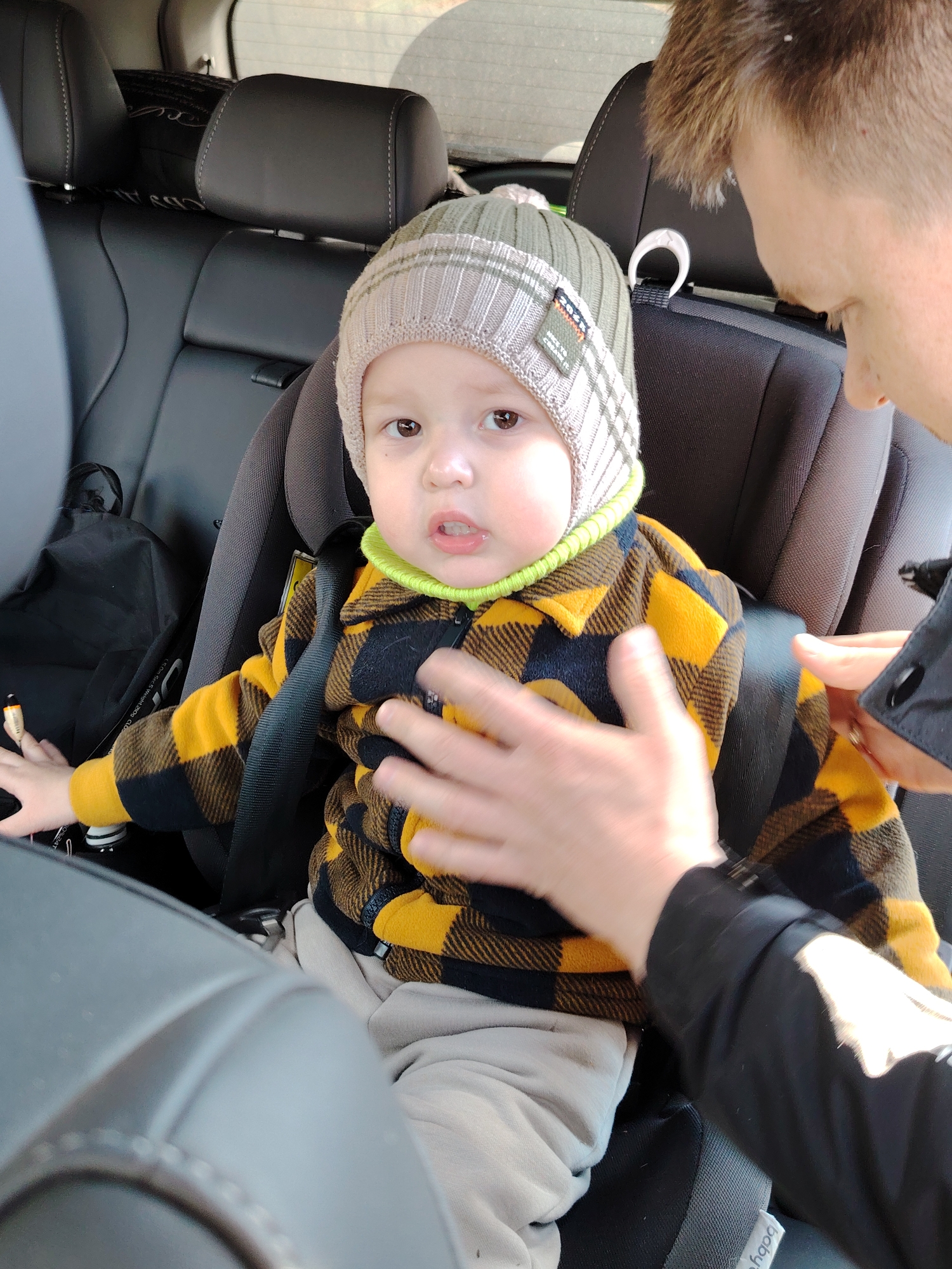A toddler in a car seat is being secured by an adult. The child is wearing a grey and white plaid hat with a green trim, a yellow and black plaid shirt, and grey pants. The car seat is black. The adult's hands are visible, and they are wearing a black jacket. The location is inside a car, likely in Chaikovsky, Russia, based on visual elements common to that region. The scene suggests the adult is preparing to drive with the child. The background shows the car's interior, including the seats and window.