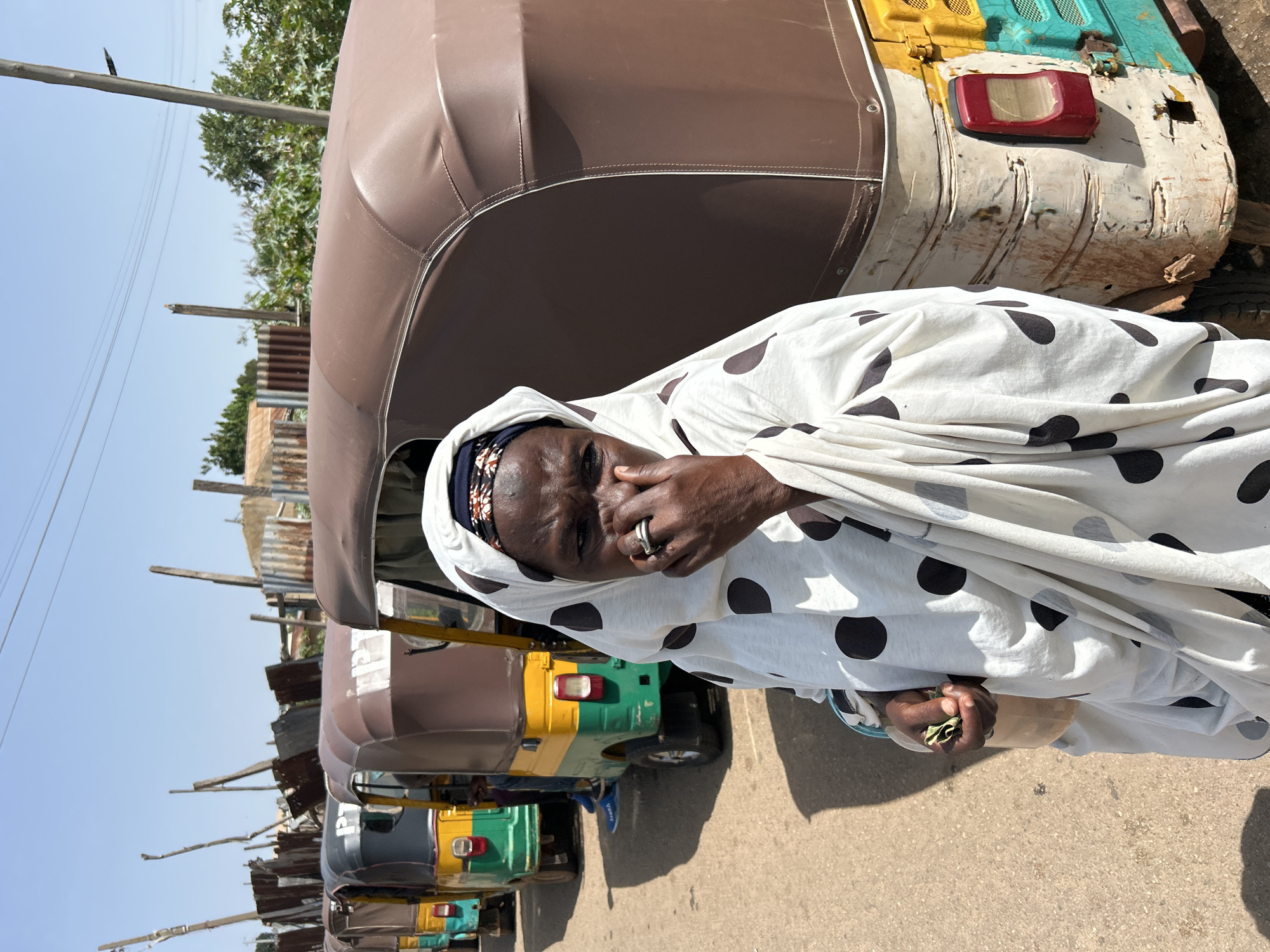 A woman, appearing to be of African descent and wearing a white headscarf with black polka dots and a dark head covering, stands in the foreground, facing the camera with her hand raised to her nose, her expression somber. Behind her, a line of colorful auto-rickshaws, commonly known as "Keke NAPEPs" in Nigeria, is parked on a dirt road. The auto-rickshaws are predominantly yellow and green with brown canopies. The environment suggests an outdoor setting, possibly a transportation hub or a marketplace in Jos, Nigeria, given the presence of the Keke NAPEPs and the style of construction in the background, which includes corrugated metal sheeting and wooden poles. The sky is clear and blue, indicating a sunny day. The overall scene depicts a moment of daily life in an urban or semi-urban environment in Nigeria.