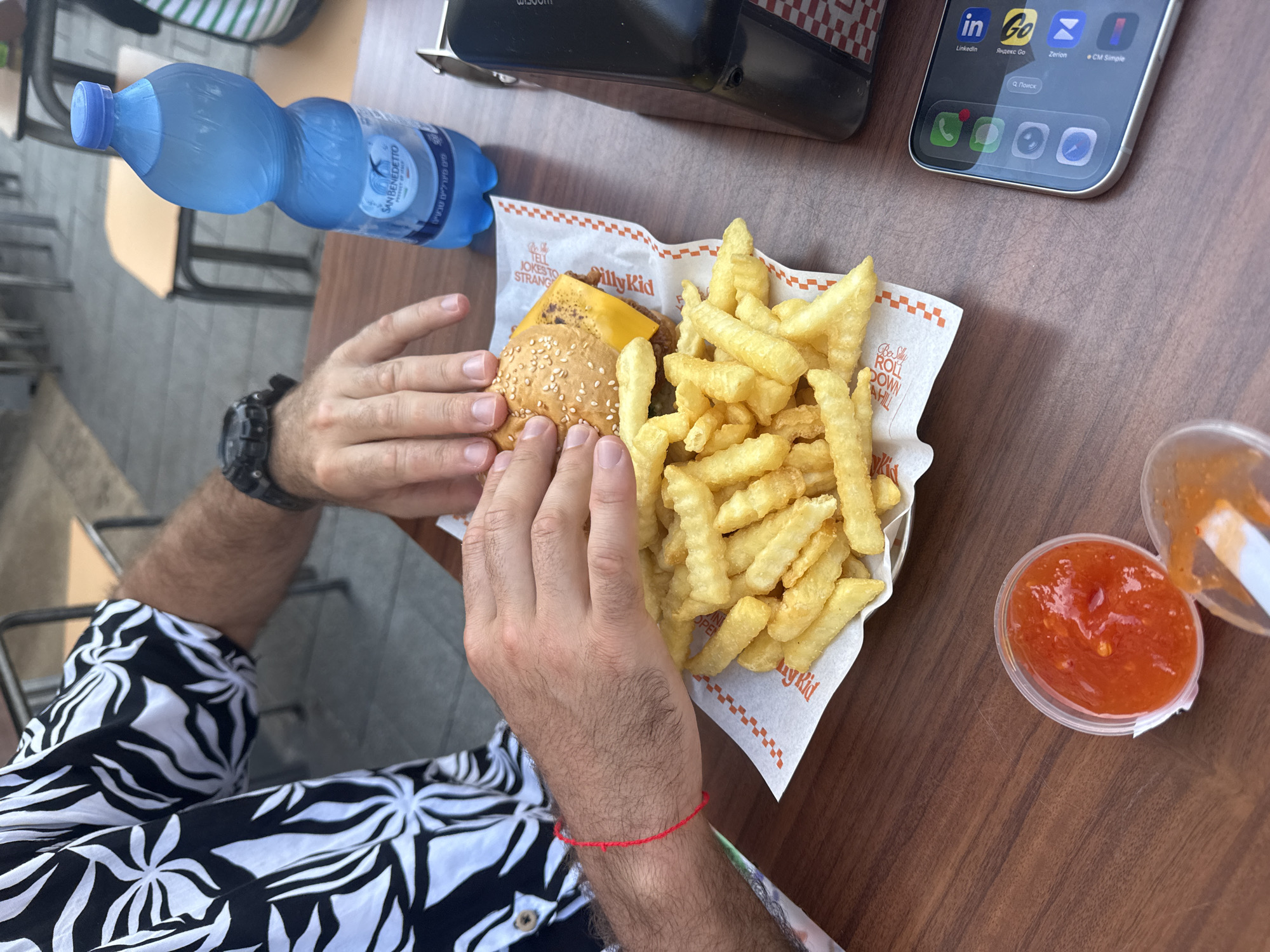 The image shows a person at a table, about to eat a burger and fries from "Silly Kid," a Tel Aviv-based fast food restaurant. Visible on the table are a bottle of San Benedetto water, a smartphone, and a container of red sauce. The person is wearing a black and white floral patterned shirt and a watch on their left wrist, and a red string bracelet on their right wrist. The table is wooden. The smartphone displays app icons for LinkedIn, Yandex Go, Zerion, and CM Simple.