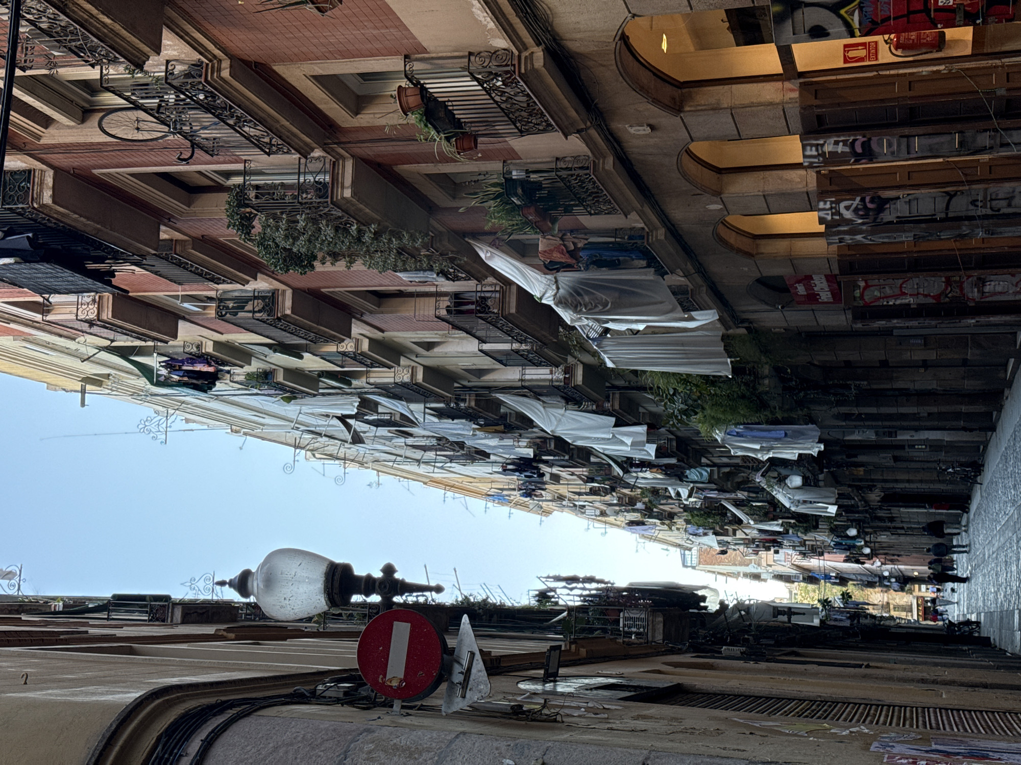 The image shows a narrow street scene in Barcelona, Spain, viewed from a low angle. Balconies with plants, hanging clothes, and awnings extend from the buildings on both sides, creating a sense of enclosure. A no-entry sign and a street lamp are prominent in the foreground. People walk along the cobblestone street, which is dimly lit compared to the sky visible between the buildings. Storefronts line one side of the street, showcasing signs and merchandise. A bicycle is mounted on one of the balconies. The overall impression is of a bustling, historic urban environment.