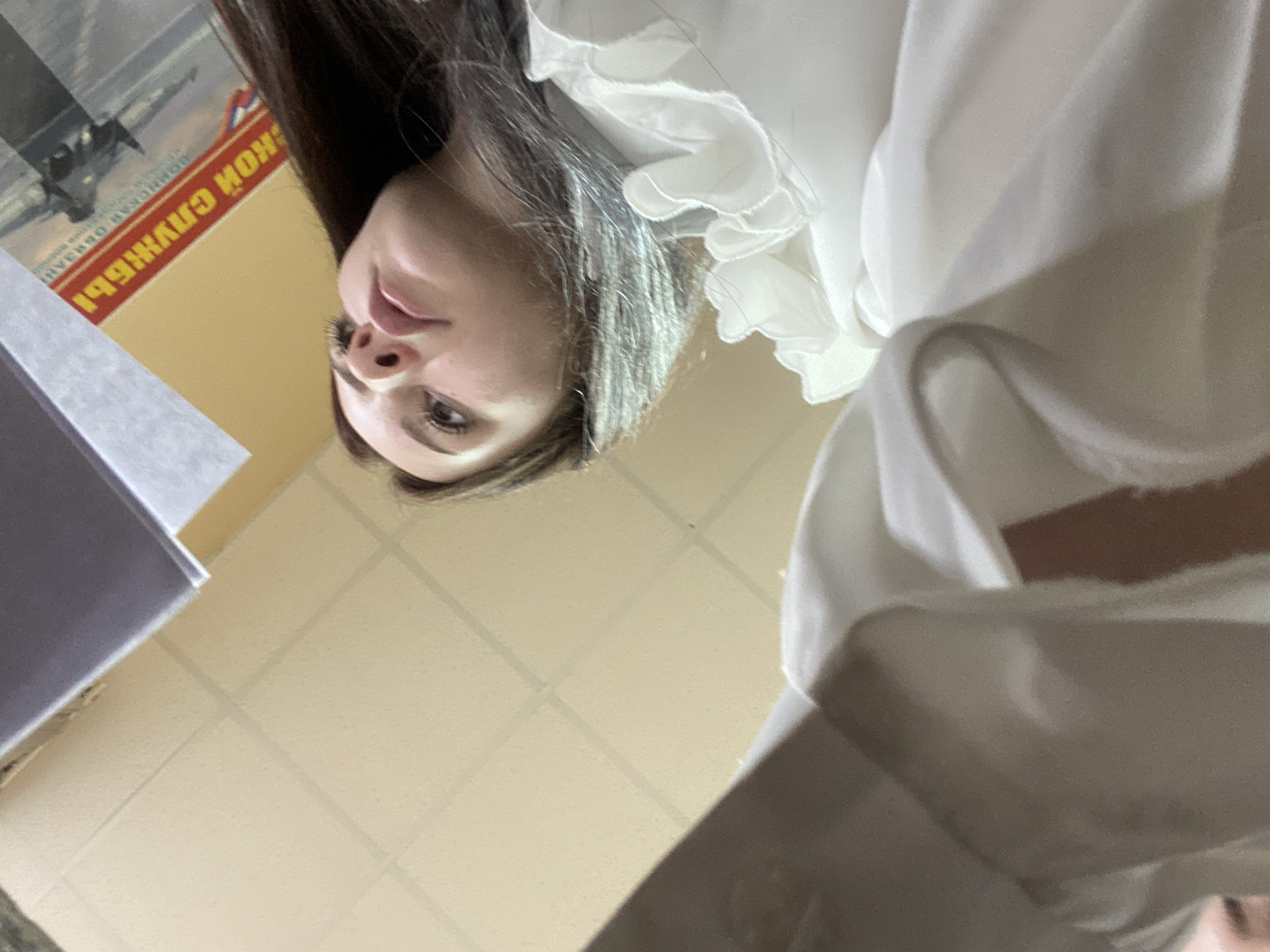 The image is an upward-facing, low-angle shot of a young woman looking away from the camera. She is wearing a white ruffled blouse. In the background, a yellow wall with a grid pattern and a partially visible red and yellow sign with Russian text are present. The sign appears to be a poster or advertisement, with the text "КОНСЛУЖБЫ" visible. The scene is indoors, possibly an office or public building, given the wall and sign. There are no indications of this location being in the city of Dyakovo, Russia.
