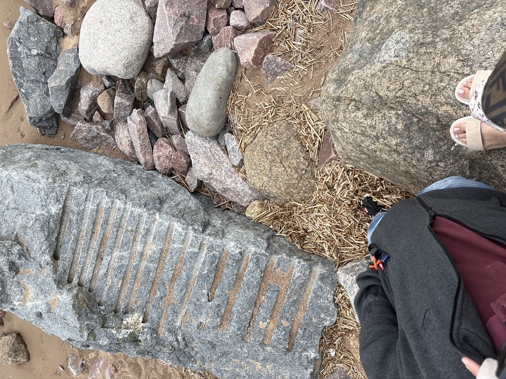The media file shows a top-down view of a rocky shoreline with sand and dried grass. In the foreground, a large, rough gray rock with carved parallel lines is prominent. To the right, the lower legs and feet of two people are visible. One person is wearing light-colored sandals and patterned leggings, standing on a large, speckled boulder. The other person is wearing blue jeans and a dark gray zip-up jacket, with a glimpse of a burgundy lining. The scene appears to be outdoors, possibly a beach or lakefront, with natural light suggesting daytime. The overall impression is casual, with people resting or exploring a natural environment. There is no visible text in the media file.