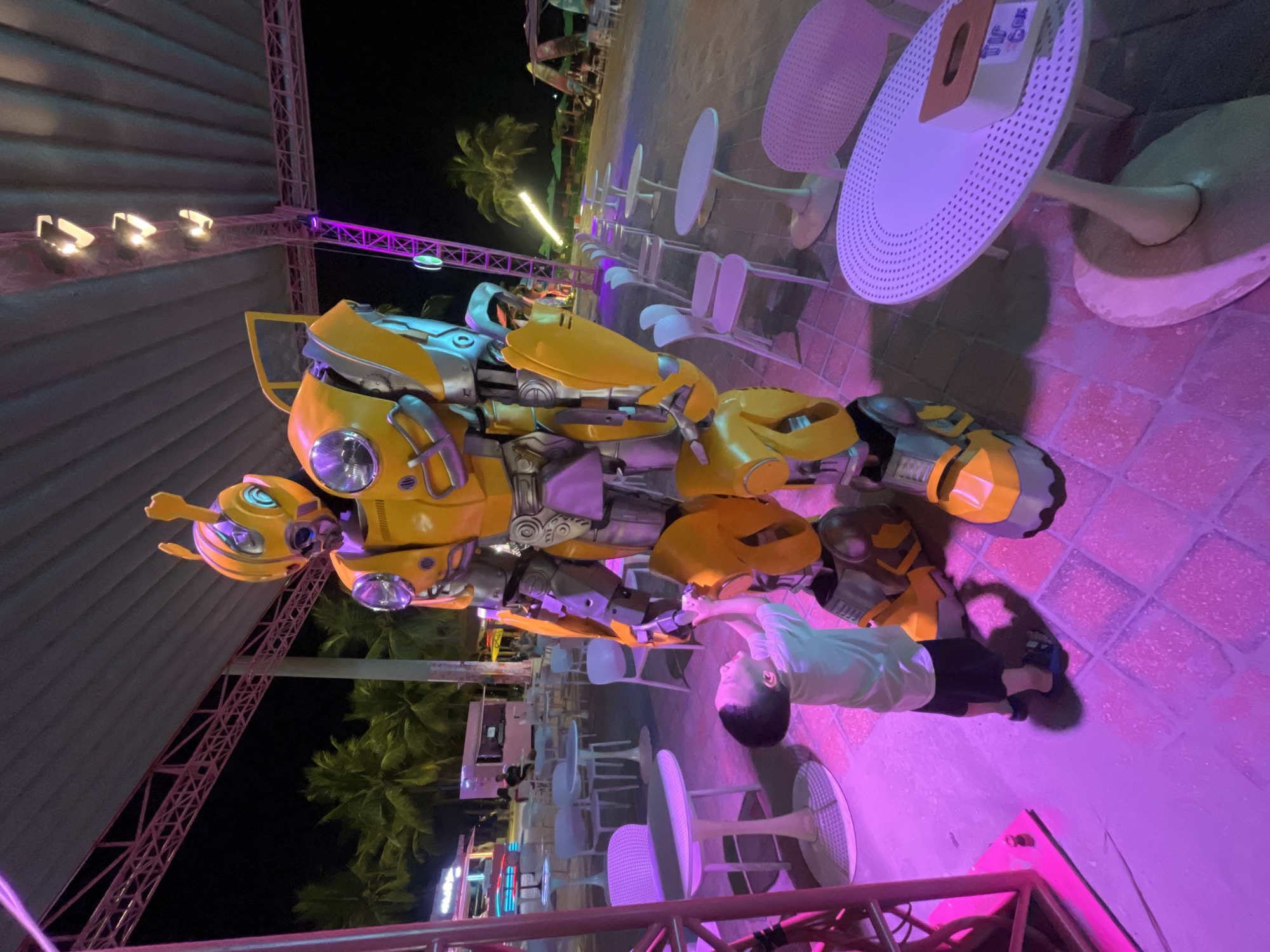 A young boy, wearing a light blue t-shirt and dark shorts, is standing on a tiled patio and reaching up to touch a large, yellow, Bumblebee Transformer costume. The costume is detailed with silver and grey accents, and its head features characteristic antennae and large, round headlights. The scene is illuminated by vibrant pink and purple lights, casting long shadows on the ground. In the background, there are several white tables and chairs arranged around the patio. Palm trees are visible beyond the covered area, and further in the distance, food stalls or small shops with neon lights can be seen. The setting appears to be an outdoor entertainment area or a restaurant at night. The image is likely taken in Đà Nẵng, Vietnam, given the presence of palm trees and the generally tropical atmosphere.
