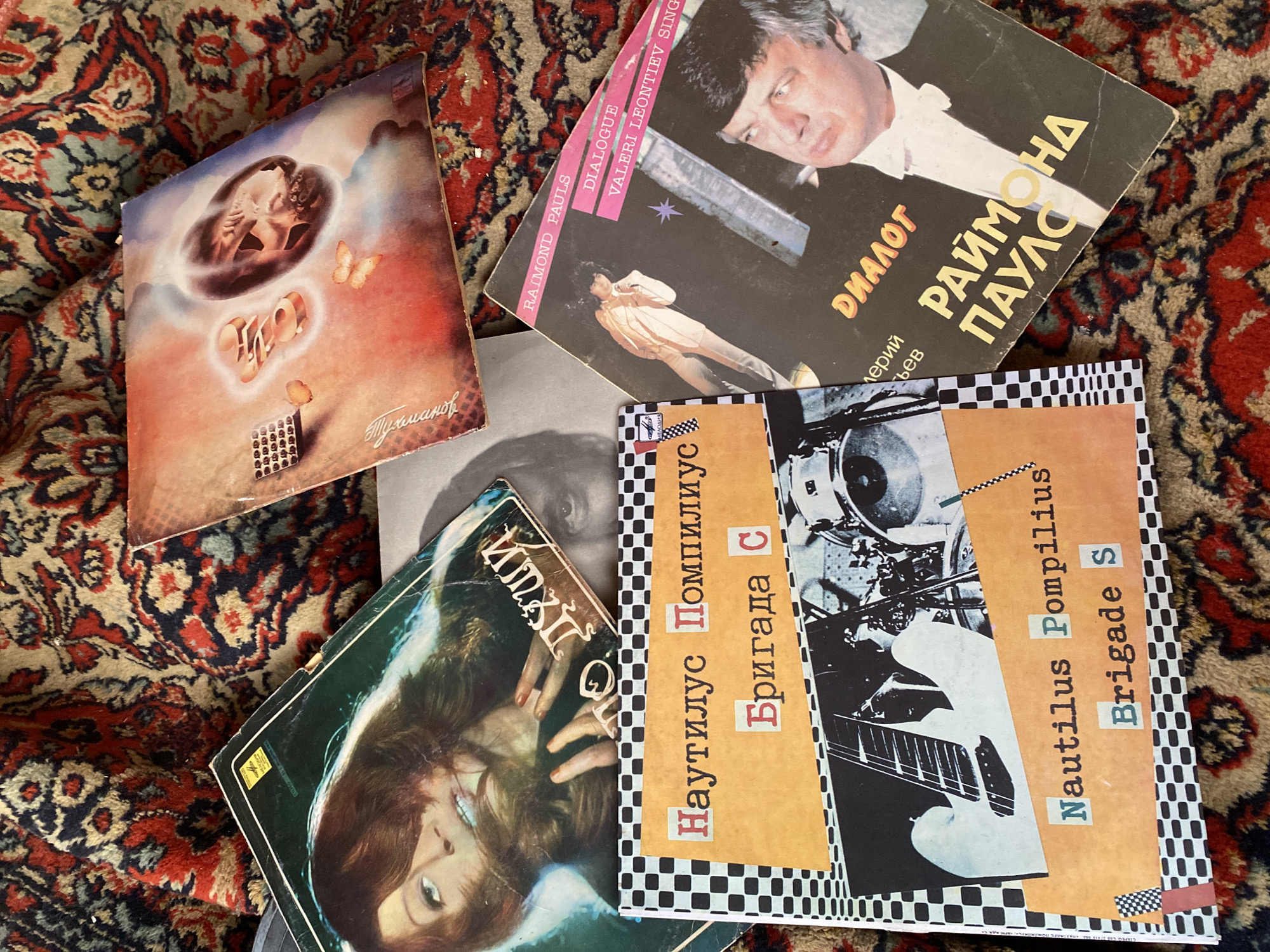 The image shows a collection of vinyl records scattered on a patterned rug. The records appear to be of Russian or Soviet-era artists and bands. Among the records are: a record featuring the name "Тухманов" ("Tukhmanov") and an image of figures in a celestial cloud, a record by "Nautilus Pompilius" with the title "Brigade S," a record by "Раймонд Паулс" ("Raymond Pauls") with the title "Диалог" ("Dialogue") featuring "Valeri Leontiev," and a record with an image of a woman with her hands near her face. There is no information in the picture about location.