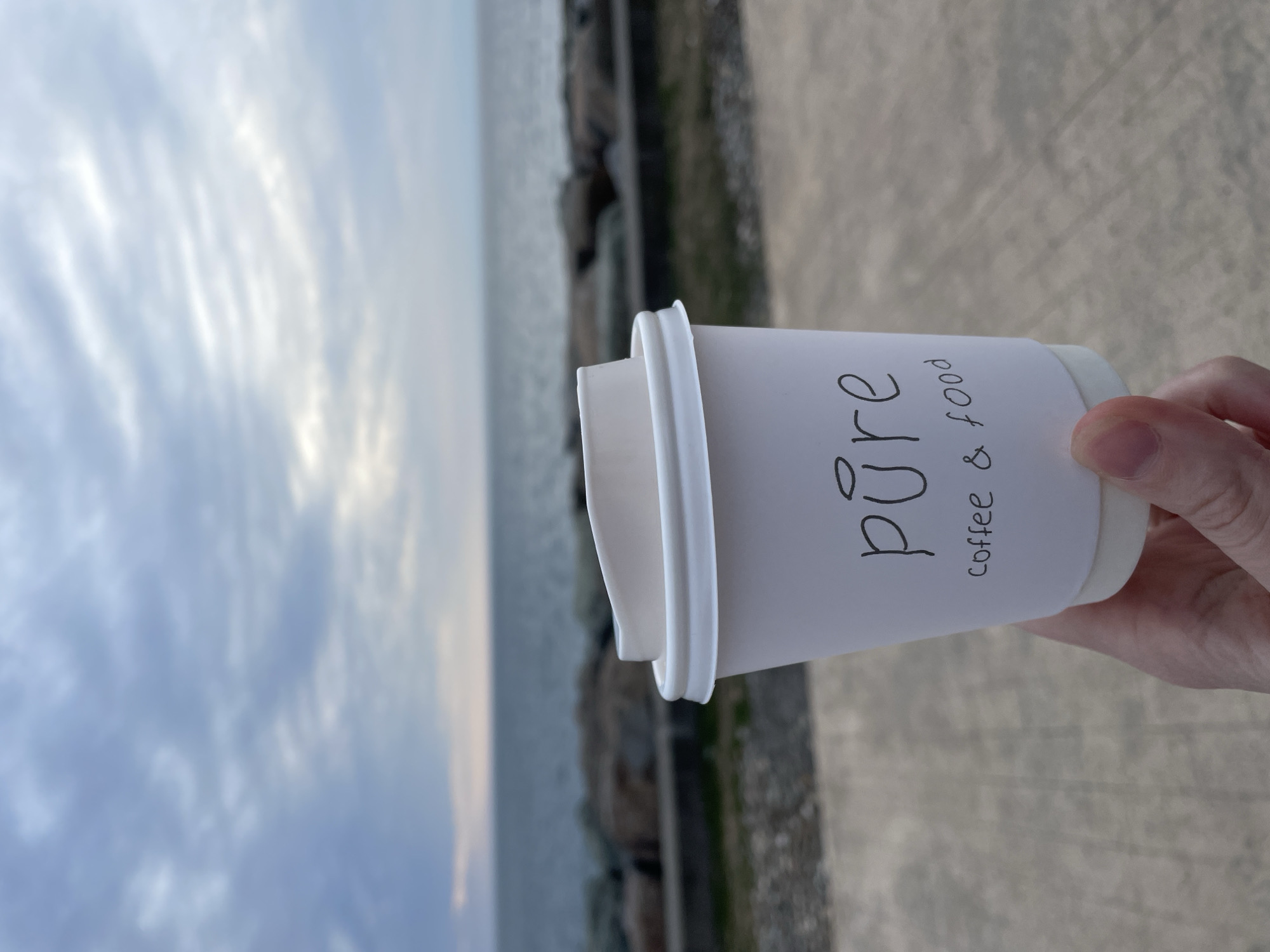 A hand holds a white paper cup with "pure" and "coffee & food" written on it. The background shows a cloudy sky, a body of water, and a rocky shoreline. The image was likely taken in Batumi, Georgia, given the presence of "Pure Coffee & Food," a cafe chain in that city. The subject is interacting with an object, the paper cup, in an outdoor environment.