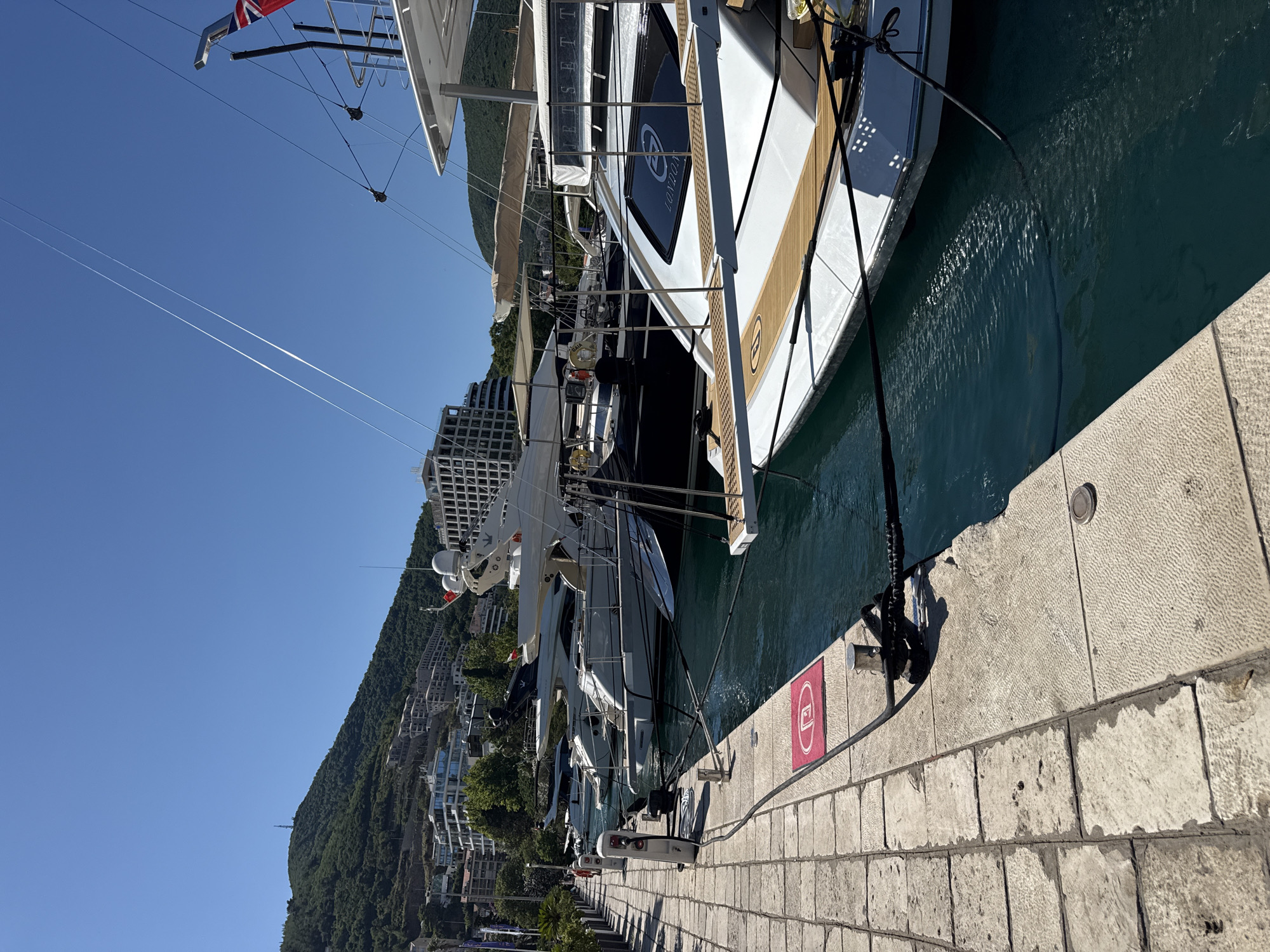 The image shows a sunny day in the marina of Budva, Montenegro. Several luxury yachts are docked along a stone promenade. The water is a clear turquoise color, and the sky is a vibrant blue with contrails from an airplane. In the background, lush green hills rise behind the city, dotted with buildings. A person is visible walking along the promenade. There are charging stations for boats along the dock. One yacht has the word "LONDON" visible on its hull.