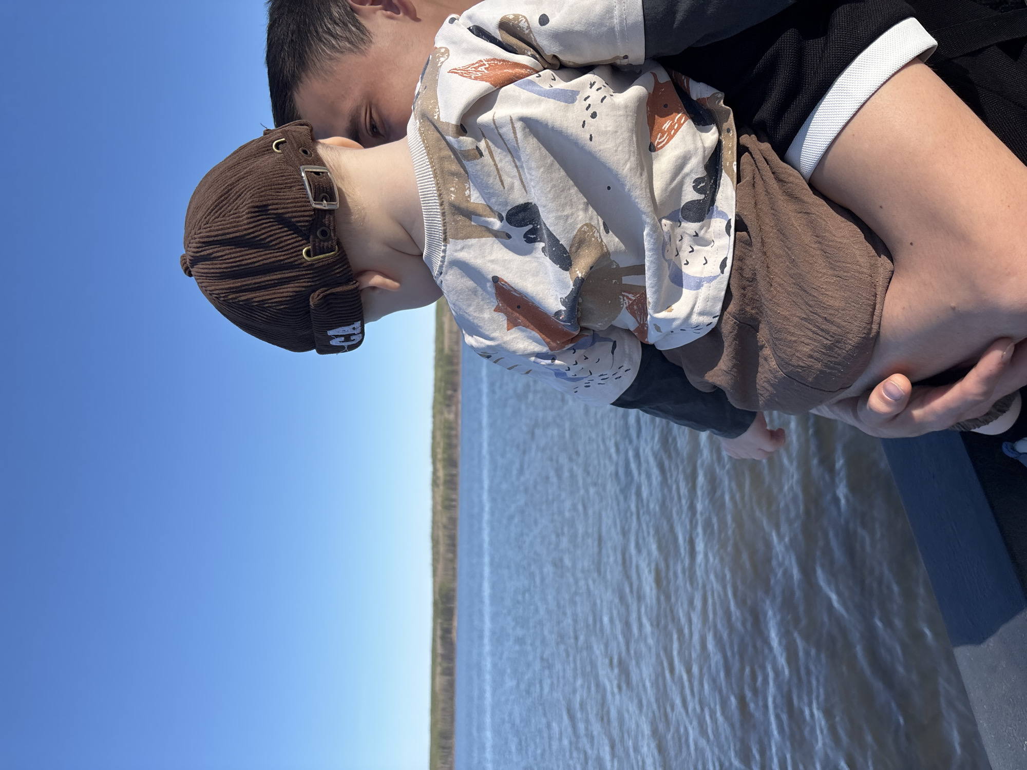 A man is holding a toddler in his arms in an outdoor scene. The man is wearing a black top with white cuffs. The child is wearing a brown cap and a patterned long-sleeved shirt. The scene includes a body of water and the sky. This photograph was taken in the city of Chaikovsky, Russia.