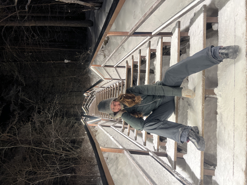 The image displays a female individual seated on the Torgashinsky Staircase in Krasnoyarsk, Russia, at night during winter conditions. The subject, with long brown hair visible beneath a grey knit beanie, wears a dark green quilted puffer jacket, dark grey winter trousers, light-colored mittens, and black boots with thick soles. She is positioned on one of the snow-dusted steps, smiling directly at the camera, with her right leg bent and left leg extended. Her right hand, covered by a mitten, rests near her jawline, and her left hand, also gloved, rests on her left thigh. The multi-tiered staircase, featuring wooden or metal steps and bronze-toned metal handrails, recedes into a dark, snow-covered forest. A continuous strip of white LED lighting is integrated along the interior edge of the handrails, illuminating the immediate area of the staircase. Snow covers the steps and the ground adjacent to the structure. Bare trees with visible branches dominate the dark background.