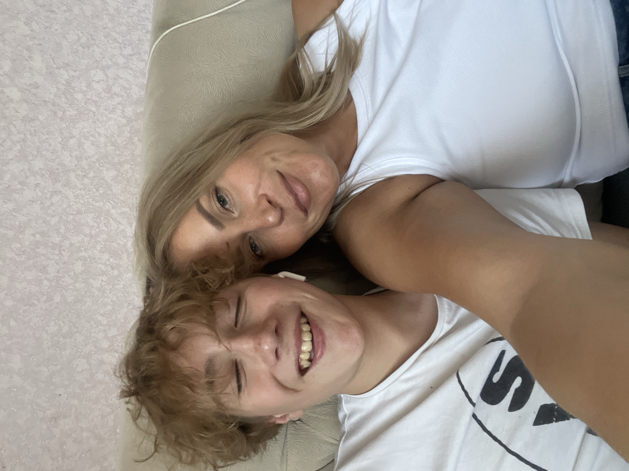 Two people are lying down next to each other. One person has blonde hair, and the other person has curly brown hair. Both people are wearing white shirts. The person with curly hair is smiling and has their eye closed. The location is not discernible from the image.