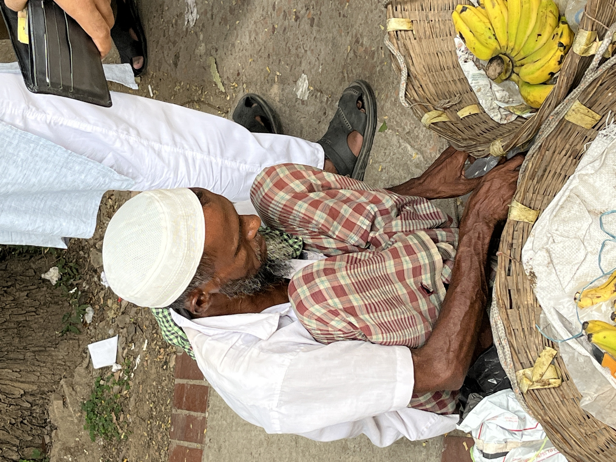 A middle-aged man, likely a street vendor, rests on the ground beside two baskets. The man is wearing a white kufi cap, a white collared shirt, and a red and brown plaid cloth wrapped around his legs. His arms are positioned around the baskets. One basket contains a bunch of ripe yellow bananas. Nearby, an individual is seen holding a black wallet, and another person's lower legs and sandals are visible. The background includes a concrete surface, possibly a sidewalk, with some dirt and debris. This scene is likely in Dhaka, Bangladesh, based on the clothing and general environment typical of South Asian urban areas.