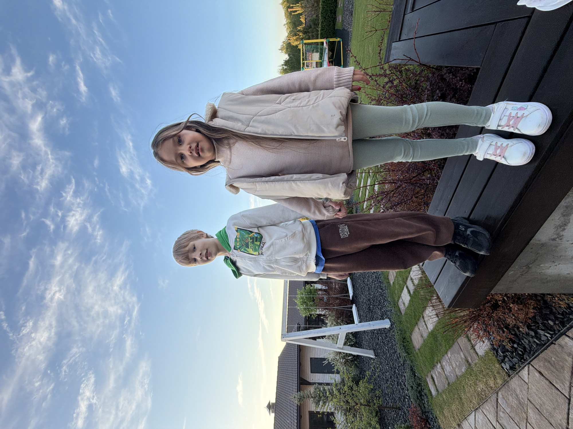 Two children, a girl and a boy, stand on a dark wooden deck outdoors. The girl, positioned above the boy in the frame, wears a light pink jacket over a lighter pink sweater, light green leggings, and white sneakers with pink accents. The boy is wearing a white zip-up hoodie with green and blue accents, brown sweatpants with lettering on the leg, and black sneakers. The background includes a blue sky with scattered white clouds, patches of grass, some bushes, and a building in the distance with a white swing set in front of it. It is not possible to determine the location based on the image.