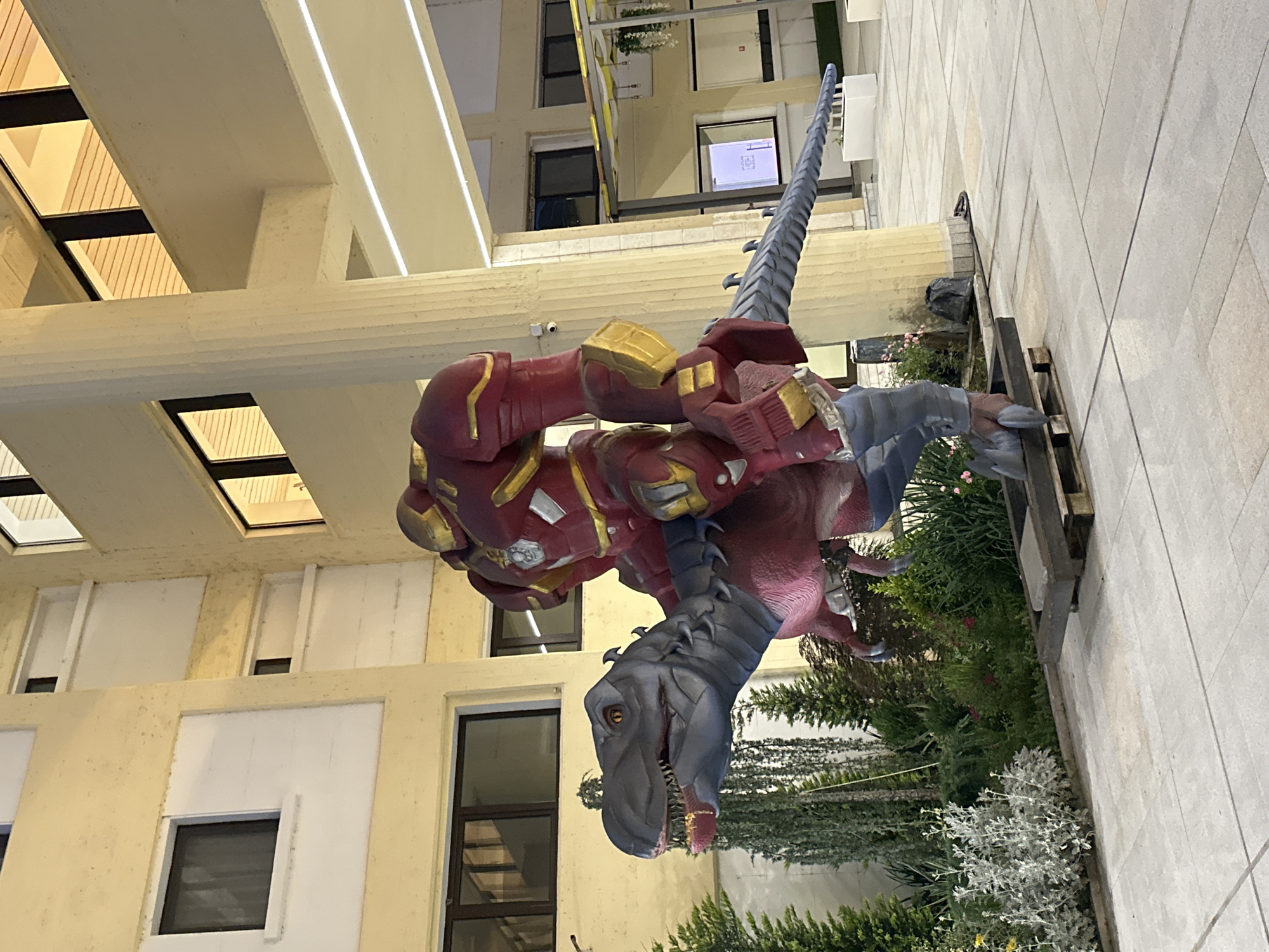 The image depicts a large, detailed statue of a dinosaur, possibly a Tyrannosaurus Rex, with a figure resembling the Hulkbuster armor from Marvel comics riding on its back. The dinosaur is primarily grey and pink, with sharp teeth visible in its open mouth and a long, spiked tail. The Hulkbuster figure is red and gold, with a robust, armored appearance. Both statues are highly realistic and appear to be made of durable materials.
The statues are positioned outdoors on a paved area, likely a plaza or walkway. Behind them, a multi-story building with a light-colored facade and numerous windows is visible. The building's architecture suggests it could be a modern office building or educational institution. There are also lush green plants and trees surrounding the statues, adding a natural element to the scene. The lighting suggests it might be evening or early morning, with artificial lights from the building illuminating the area.
The image does not contain any information about the city of Veliko Tarnovo, Bulgaria, or any specific landmarks that would place the scene in that location.