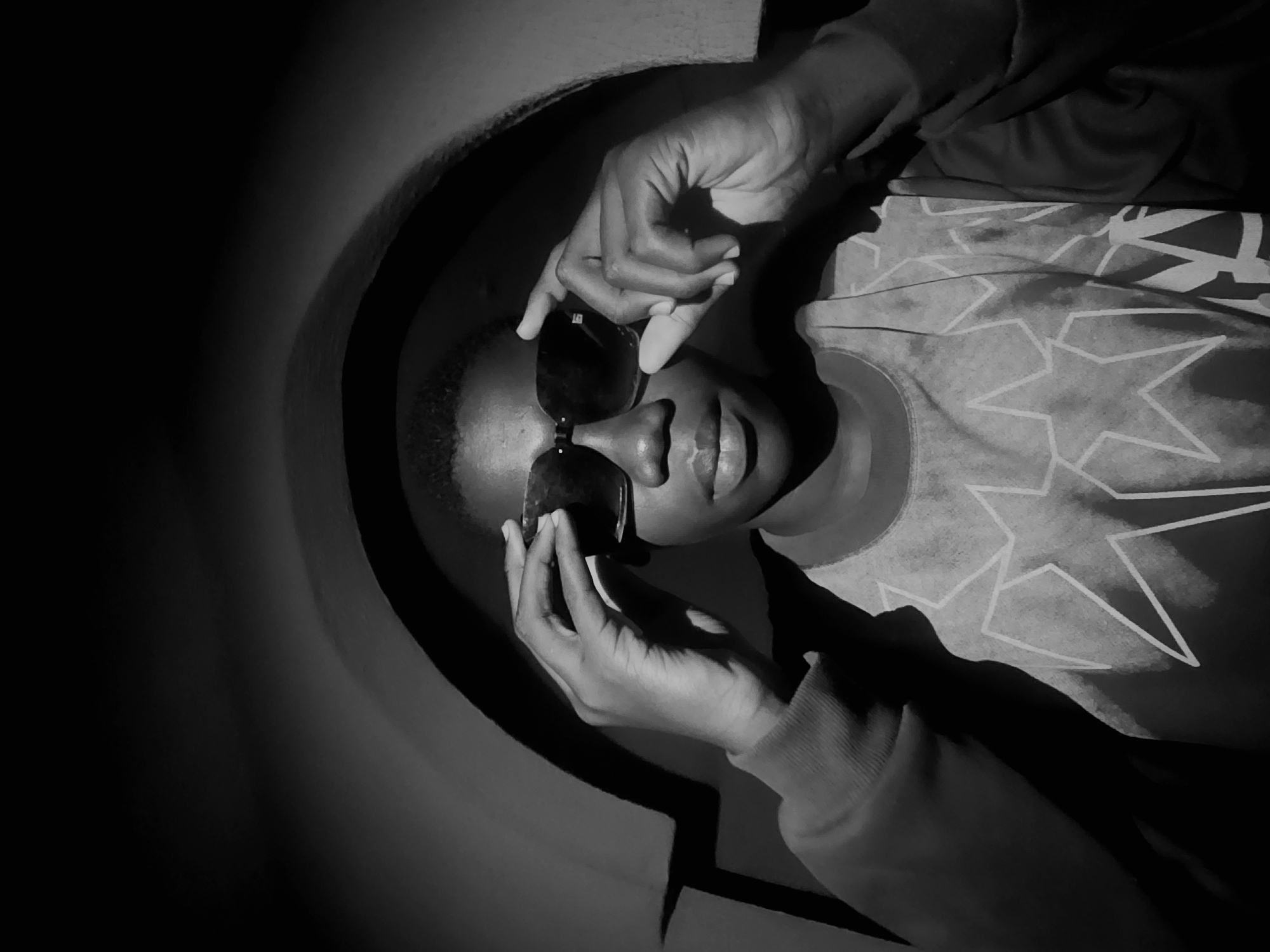 A black and white, high-angle, close-up shot depicts a person wearing sunglasses in what appears to be a round, enclosed structure. The person's face is visible, with hands holding the sunglasses. The person is wearing a long-sleeved top with a star pattern. The surrounding environment is mostly dark, with the light focused on the person and part of the circular structure. There are no obvious identifiers of a specific location in Asaba, Nigeria.
