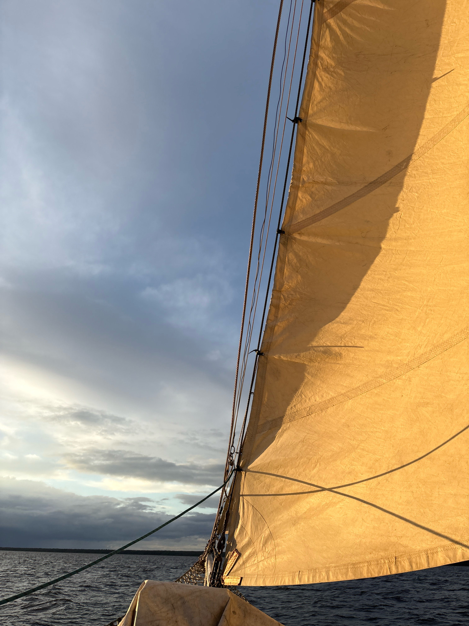 This is an outdoor, close-up, eye-level shot of a sailboat on the water, viewed from the perspective of someone on the boat. The main subject is a large, tan sail, brightly lit by the sun on its right side, with a shadow cast across its surface. The sail is taut and billows slightly. Several ropes are visible running vertically along the left edge of the sail, attached to the mast. Beyond the sail, the sky is a mix of blue and gray clouds, with a patch of lighter sky peeking through on the left. The water is dark blue and choppy, with small waves. In the distance, a dark, tree-lined horizon is visible. The image appears to be taken during either sunrise or sunset, given the warm light on the sail and the dramatic clouds. There are no people or animals visible in the image, and the location is not Saint Petersburg, Russia.