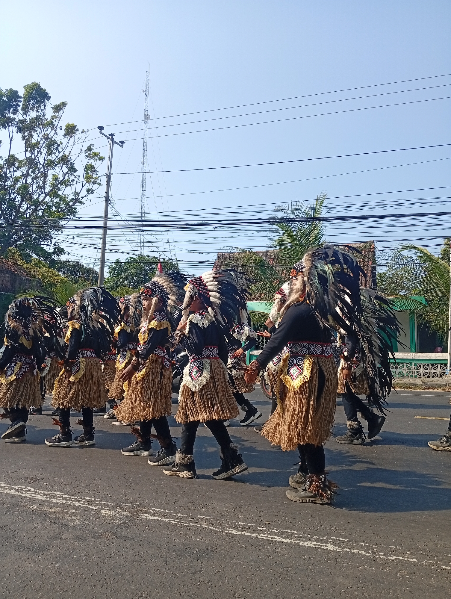 The image shows a group of people in traditional attire marching in a street in Sarang, Indonesia. The individuals are wearing feathered headdresses, black tops, and brown fringed skirts. They are walking on a paved road with white lines marking lanes. In the background, there are trees, houses, power lines, and a tall tower structure. The sky is clear and blue. The people appear to be participating in a parade or cultural event.