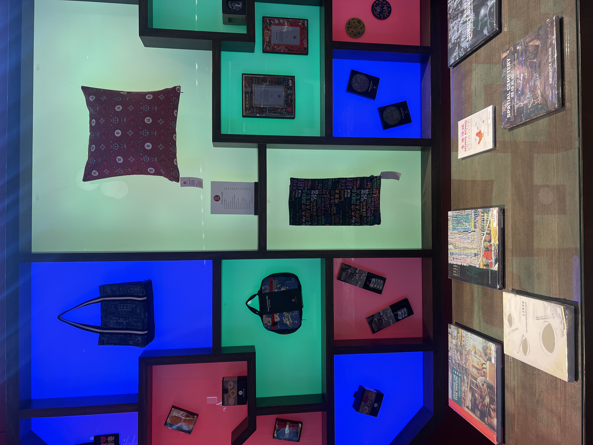 This image depicts a retail display within what appears to be a museum or gift shop in Hong Kong, China. The display is arranged in a grid of brightly colored illuminated cubbies against a dark background.
In the upper left cubby, a dark fabric tote bag with white straps is displayed against a blue background. Below this, a small red cubby contains a small package.
To the right of the tote bag, in a larger green illuminated cubby, a red patterned cushion with a tag is suspended by wires.
Further down, in a green cubby, a blue patterned travel toiletry bag is hanging. To its right, a framed certificate or document is displayed in a green cubby, and another framed piece is in a red cubby.
In a blue cubby on the lower left, a small black box is displayed. Below this, in a pink cubby, two rectangular items with text and graphics are arranged vertically.
In a central green illuminated cubby, a black fabric item with white text is displayed vertically, with a tag hanging from it.
At the bottom of the display, on a wooden surface, several books are arranged. From left to right, these include a book titled "DRAWING ON THE INSIDE," another titled "HONG KONG," a white book with Chinese characters and the title "GRANDMA GRANDMA," a book titled "HONG KONG" with a small orange graphic, and a book titled "SPATIAL CEMETERY A JOURNEY THROUGH URBAN LANDSCAPES OF HONG KONG." To the far right, a book with a grayscale cover is partially visible, possibly titled "SUNSET."
The overall scene suggests a curated exhibition of goods, potentially souvenirs or art pieces, related to Hong Kong culture and landmarks. The use of vibrant colors and the arrangement of items indicate a deliberate design for visual appeal and to highlight the products. There are no people visible in the image.