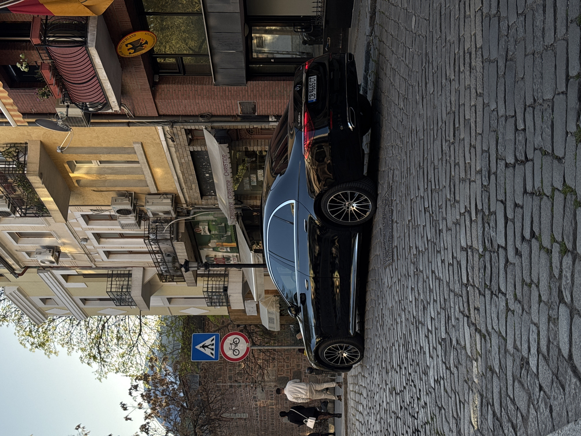 The image shows a street scene in Plovdiv, Bulgaria. The street is paved with cobblestones and has buildings on both sides. A black Mercedes-Benz S400d is parked on the street, its license plate reading "CB 9665 CT". A pedestrian crossing sign and a "no bicycles" sign are visible near the front of the car. Two people are walking on the sidewalk near the car. Above the building on the right there is a sign that reads "Deutschland".