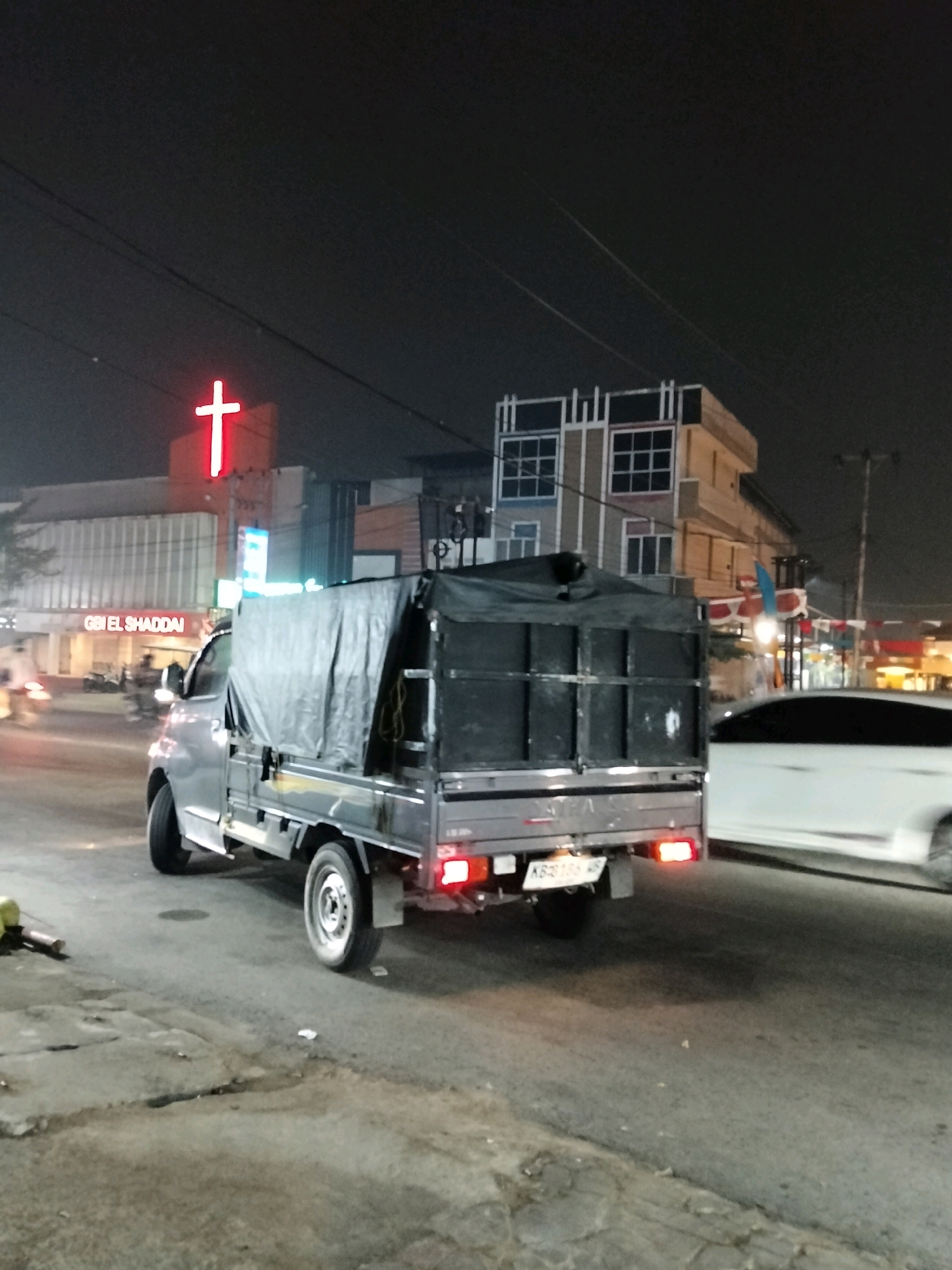 This is a nighttime, eye-level, medium shot of a street scene in Pontianak, Indonesia. In the foreground, a grey pickup truck with a black tarp covering its cargo bed is driving away from the camera, facing right. The truck has its brake lights illuminated. To the right of the truck, a white car is driving in the same direction. In the background, a building with a prominent red neon cross sign is visible. Below the cross, the text "G BI EL SHADDAI" is illuminated in white. There are other buildings and streetlights in the background, suggesting an urban environment. The road surface is asphalt and appears wet in places.