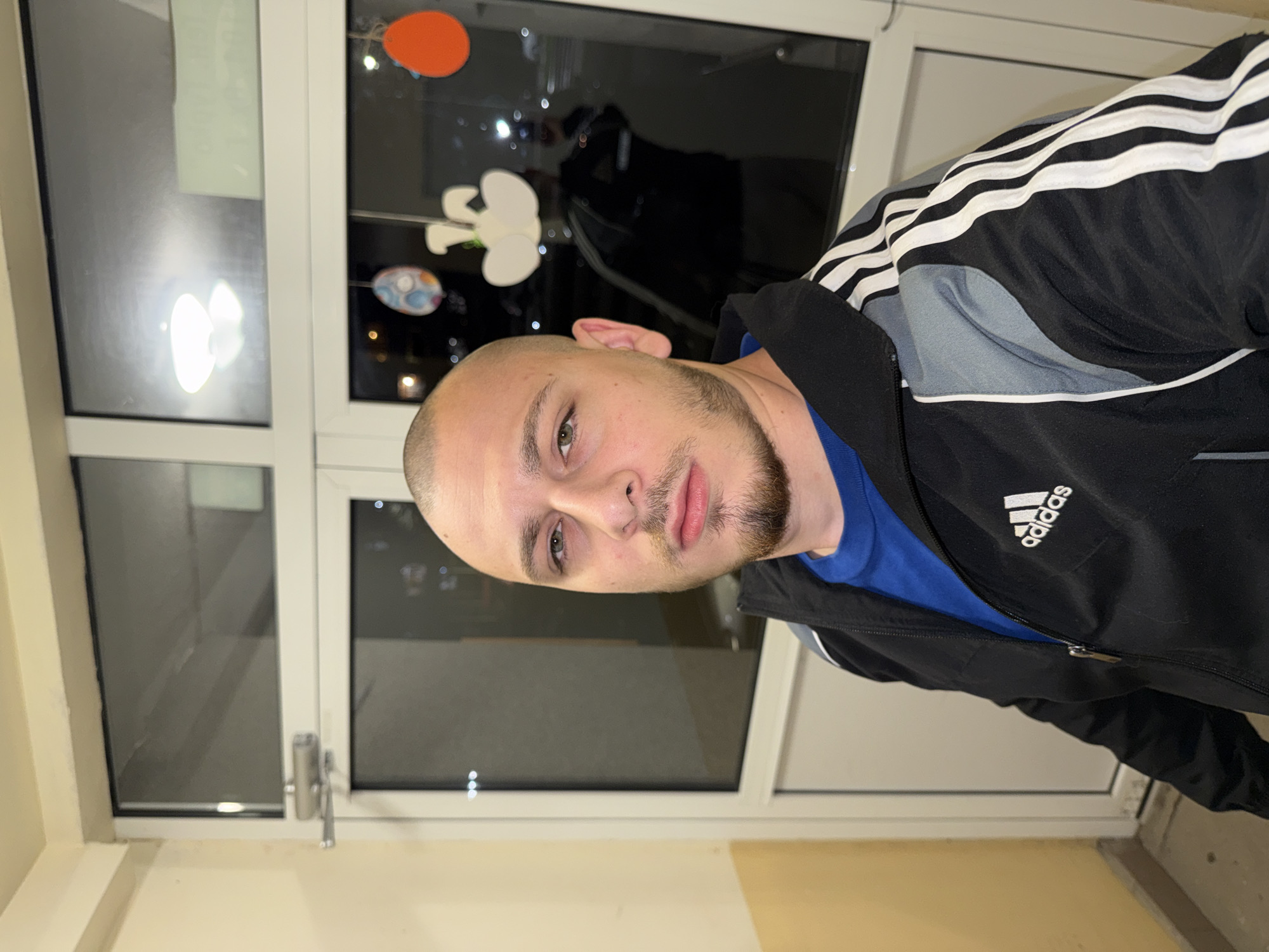 A medium-height, light-skinned man with a shaved head, stubble, and a blue shirt is wearing a black Adidas jacket with three white stripes along the shoulders and sleeves. He is looking directly at the camera. He is standing in front of a white-framed window with multiple panes. Through the window, there are reflections, including an orange circle and a white, abstract, possibly clover-shaped object. The bottom of the frame shows a light-colored wall, likely inside a building. There is no indication as to whether this location is in Stara Zagora, Bulgaria.