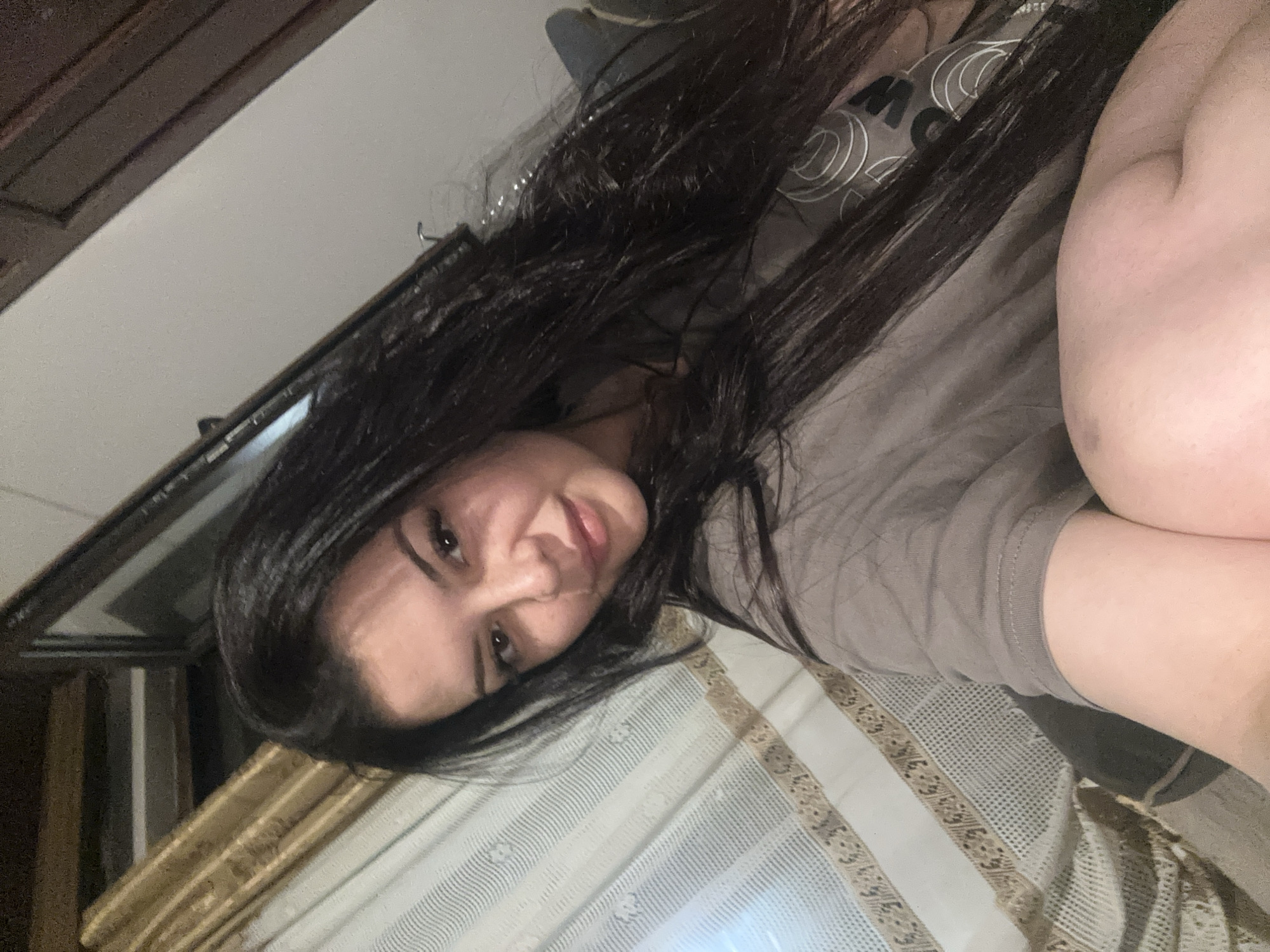 An indoor portrait taken in Chubra, Bulgaria, features a woman captured in a close-up, reclined pose. She is positioned towards the right of the frame, with her head slightly tilted. She has long, dark brown or black hair that falls over her shoulders and chest. Her dark eyes are open, and she offers a soft, pleasant smile directly to the camera, conveying a relaxed demeanor.
The woman is wearing a grey t-shirt with white or light grey circular patterns visible on the left shoulder and chest area, along with a stylized letter "M" as part of a logo or design. Her right arm and shoulder, showing bare skin, are visible in the foreground.
The background elements suggest a domestic setting. In the upper left portion of the image, dark brown wooden furniture or architectural details are visible. A light-colored wall or ceiling occupies the upper central and right areas. Beneath the woman's head and extending across the lower half of the frame is a light-colored fabric, possibly a bedspread or blanket, featuring horizontal stripes and ornate patterns along its borders. A patterned curtain or blind is also discernible in the lower left. The lighting is consistent with an indoor environment, possibly using artificial light.