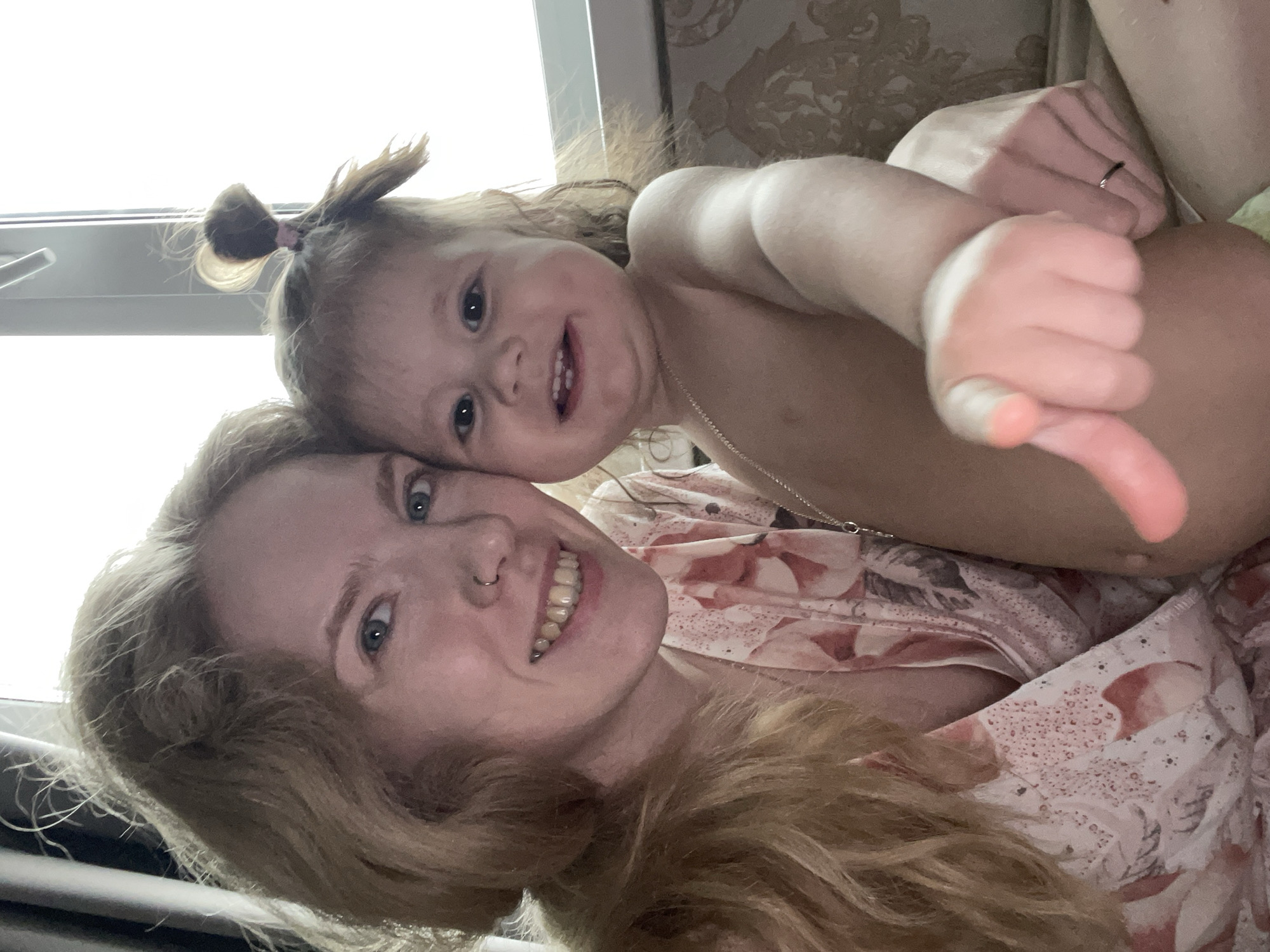 A woman with long, reddish-blond hair is lying down and smiling into the camera. She has blue eyes and a nose ring. She is wearing a light-colored floral print top. A young child is lying on top of her, also smiling at the camera. The child has light brown hair tied up in a small ponytail and is partially nude. The child is giving a thumbs-down gesture with her right hand. A silver necklace is visible on the child's chest. The background appears to be an interior setting, possibly a home, with a window visible in the top left corner. There is decorative wallpaper behind them. There is no verifiable evidence to determine the location as Izhevsk, Russia.
