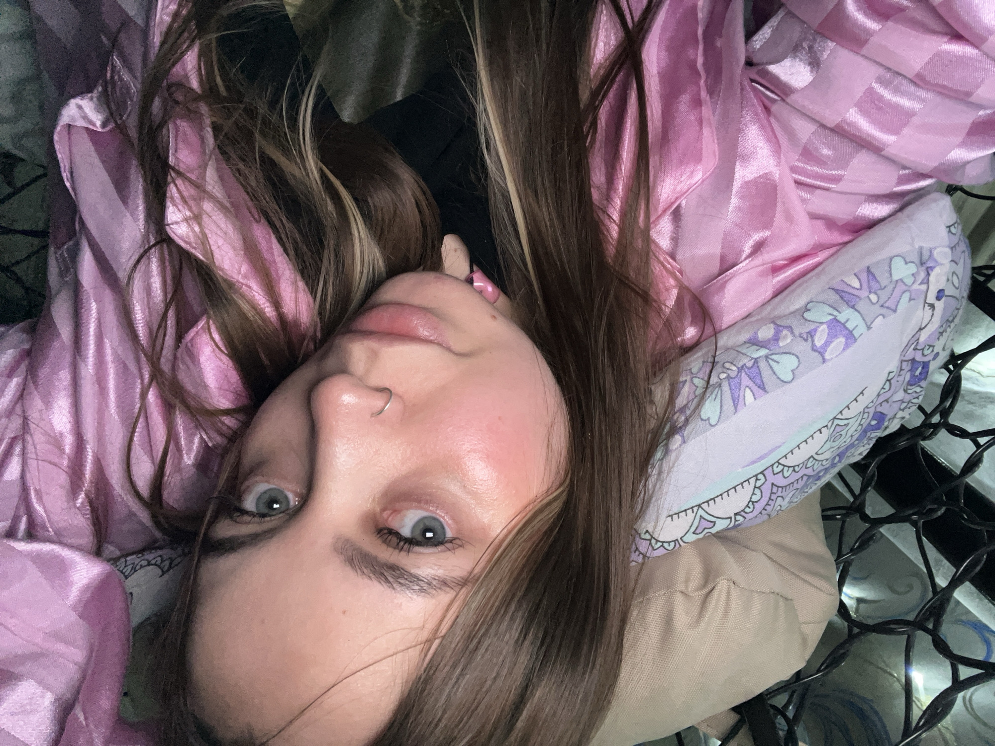 A woman with fair skin and light gray eyes is lying on her back, her head resting on a patterned pillow. She has brown hair with blonde highlights, and is wearing a pink, striped pajama top. A small heart-shaped nose ring is visible in her left nostril. The background includes a dark, possibly cluttered space, and a string of lights is visible on the right side of the frame. There is no visual information indicating a location in the city of Novopokrovskaya, Russia.