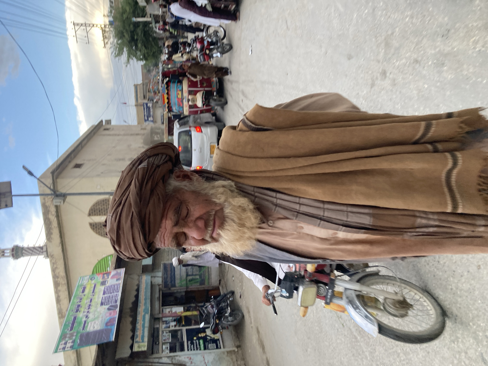 The image captures a vibrant street scene, likely in Loralai, Pakistan, featuring an elderly man on a motorcycle. The man, positioned prominently in the foreground, has a long, light-colored beard and a weathered complexion. He wears a brown turban, a large brown shawl draped over his shoulders, a light-colored shirt, and a dark vest. He appears to be smiling or smirking gently, looking directly towards the viewer while holding the handlebars of his red and silver motorcycle.
The background depicts a busy street lined with buildings and bustling with activity. To the left, shopfronts are visible, with one featuring a large green and blue sign hanging above, which appears to advertise solar panels. Another shop displays merchandise. A smaller black motorcycle is parked nearby. In the mid-ground and further down the street, various vehicles including a white car, a colorful auto-rickshaw, and other motorcycles are present, alongside several people.
The sky, visible in the upper left, is partly cloudy with bright sunlight breaking through, suggesting either early morning or late afternoon. The overall atmosphere is lively, characteristic of a local market or main street. The image is a candid shot, capturing a moment of everyday life. Any visible text on signs appears to be in a local script, likely Urdu.