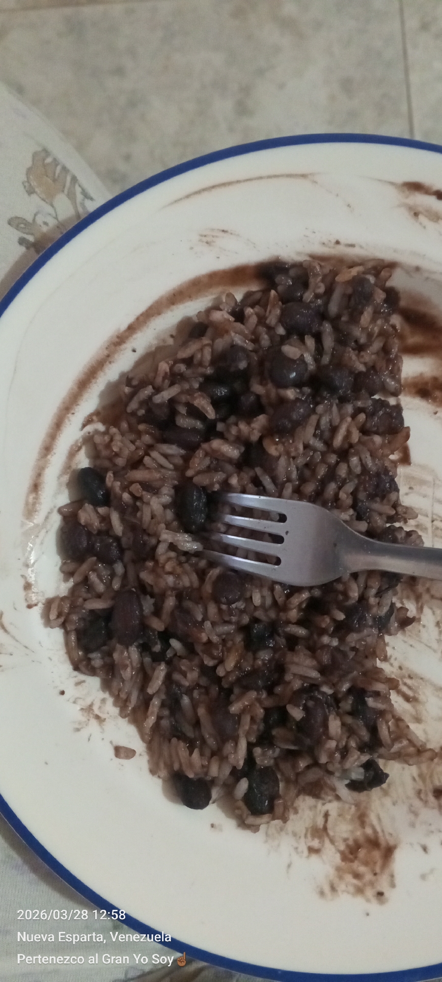 A plate of rice and beans with a fork is seen in the image. The plate is mostly eaten with a dark sauce remaining on the plate. The lower left of the image contains the date, time, and location of the image: 2026/03/28 12:58, Nueva Esparta, Venezuela. The location is not in Altagracia, Venezuela.