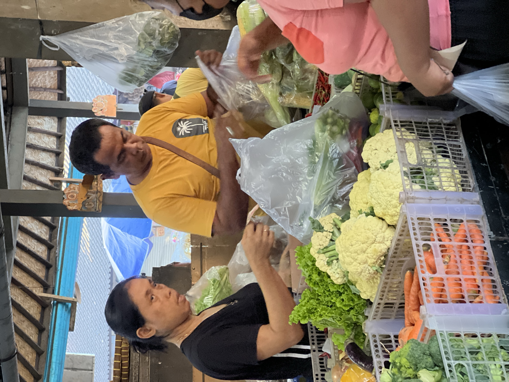 The image depicts a lively scene at a fresh produce market, likely in Ubud District, Indonesia. The setting appears to be a covered market stall, with wooden beams and a patterned woven structure visible in the background, along with a glimpse of a blue tarp or umbrella.
In the center-left, a man with short dark hair, wearing a yellow t-shirt with a black palm tree logo, is actively handling clear plastic bags filled with vegetables, suggesting he is a vendor preparing items for sale. He is looking down at the produce. To his lower left, a woman with dark hair pulled back, dressed in a black t-shirt, is looking towards the vegetables or the man, possibly a customer making a selection.
On the right side of the frame, partially obscured, another person in a pink top is also involved in handling plastic bags and produce, likely another vendor or customer. The market stall is abundant with fresh vegetables. White plastic crates stacked on the right display large, pale yellow cauliflower heads, vibrant green lettuce, and rows of bright orange carrots. Below these, additional vegetables like green broccoli and dark purple eggplants can be seen. Many other green leafy vegetables are visible, some already packed in clear plastic bags.
The activity clearly shows people engaged in buying and selling fresh produce. The lighting indicates it is daytime, with ample natural light illuminating the scene. No discernible weather conditions are visible. Aside from the palm tree logo on the man's shirt, no other specific text is clearly legible in the image.