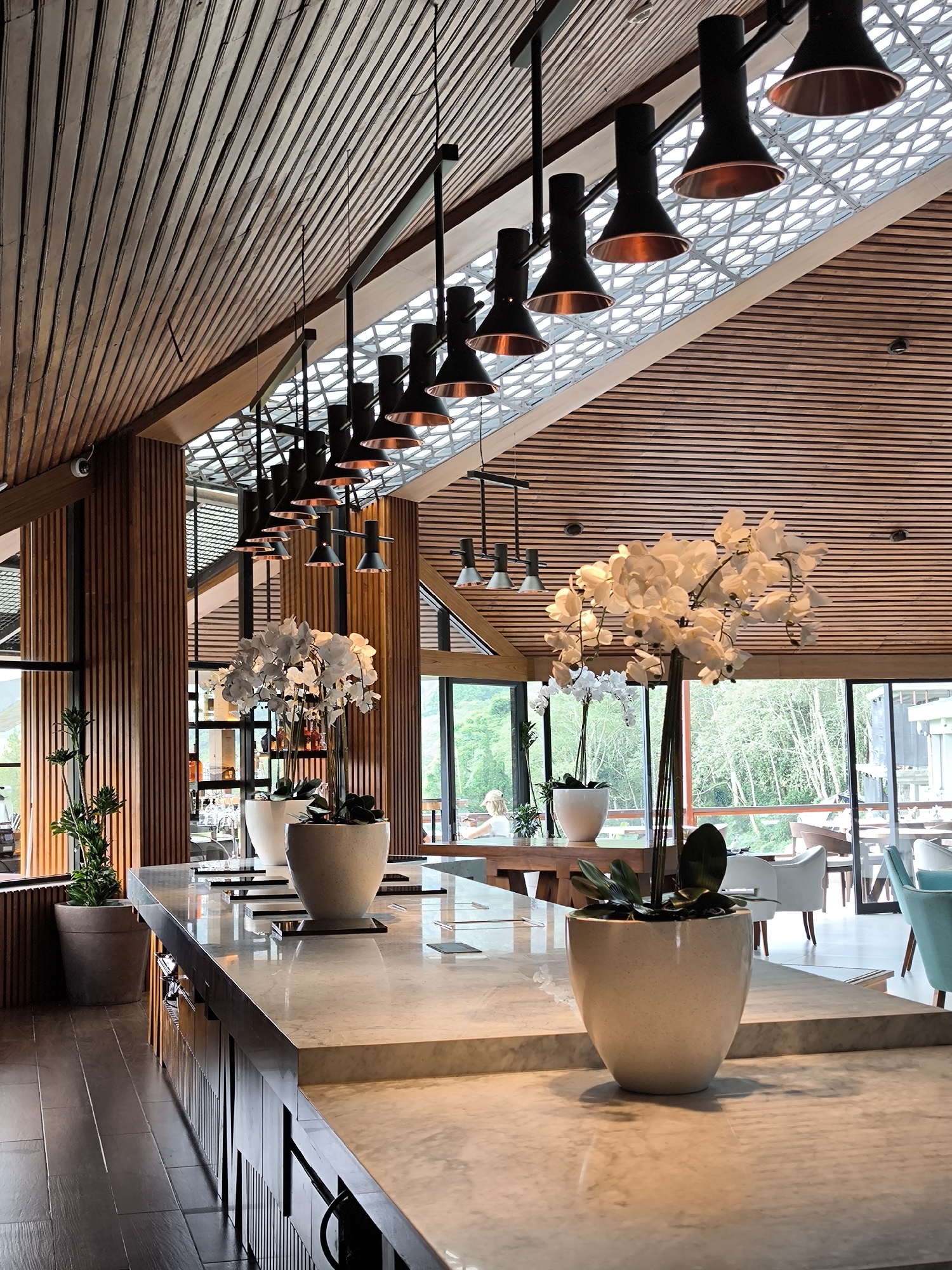 The image captures the interior of a high-end venue, potentially a restaurant or hotel lounge, located in Wanagiri, Indonesia, as suggested by the architectural design elements and natural scenery visible through the windows.
The focal point of the image includes a series of potted white orchid plants arranged along a marble countertop, likely a bar or serving area. The countertop has a glossy, reflective surface. Behind the orchids, large windows reveal a lush, green landscape. Further in the background, there are tables and chairs arranged for patrons.
Overhead, a series of black pendant lights with copper-toned interiors are suspended from the wooden ceiling. The ceiling itself is paneled with linear wooden slats, arranged in parallel lines and interrupted by the geometrical design of the skylight structure.
A portion of a wooden-slatted wall is visible on the left of the image, and adjacent to it is a potted tree.
There are no people in clear focus in the foreground, but a person is sitting at a table in the background. The general atmosphere appears upscale and serene. The lighting is soft and warm, highlighting the natural materials and colors in the scene.