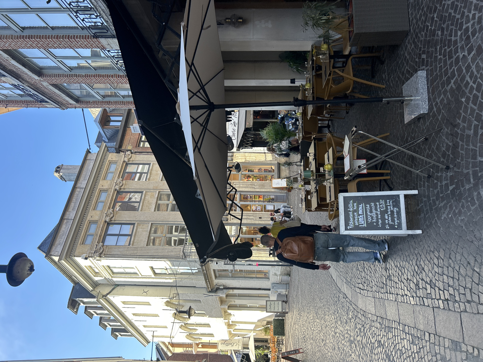 The image depicts a street scene in Leuven, Belgium, featuring the facade of a restaurant named "Restaurant Fellini" and an outdoor seating area. In the foreground, a man with a brown backpack is walking away from the camera down a cobblestone street. In front of the restaurant, several tables are set with place settings and yellow accents. A chalk sign outside the restaurant displays a lunch menu in Dutch and English, mentioning "Vegan friendly" options.
The architecture of the buildings lining the street is characteristic of European cities, with a mix of brick and stone facades, arched windows, and decorative elements. The sky is clear and blue, indicating a sunny day. The outdoor seating area is partially covered by a large black awning and a light-colored umbrella. Various potted plants add greenery to the scene. In the background, other people are visible, some seated at tables and others walking. The overall impression is that of a pleasant afternoon in a historic European city center.