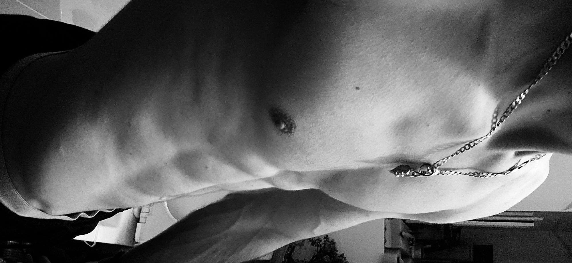 The image depicts a close-up, black and white shot of a torso, likely male, displaying defined abdominal muscles and chest. A thin chain necklace adorns the neck and upper chest area. The scene appears to be indoors, with blurred background elements suggesting a room setting. There is no information available to determine if this image was taken in Plovdiv, Bulgaria.