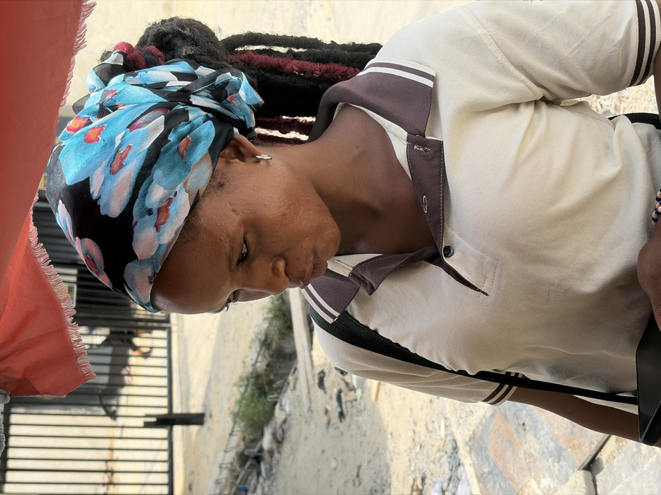 An adult female is depicted in profile, facing left, in an outdoor setting in Igboefon, Nigeria. She has dark skin and wears a black headwrap adorned with blue and reddish-pink floral patterns. Dark hair, appearing as dreadlocks with some reddish-pink strands, extends from beneath the headwrap. A single silver hoop earring is visible on her right ear. Her mouth is slightly pursed, and her gaze is directed downward and to her left. She wears a light beige short-sleeved polo shirt with a dark brown collar, white stripes on the collar and sleeve cuffs, and a visible dark button on the placket. A black strap, likely from a bag, crosses her right shoulder. Her right hand, in the lower frame, holds a dark-colored smartphone, and her right wrist bears a beaded bracelet containing blue, white, and brown elements. The immediate foreground shows a partially visible, frayed orange-red fabric canopy. The background consists of an unpaved ground surface with dirt and gravel, a shallow drainage channel to the left, and a dark metal gate or fence structure partially visible behind the subject. The scene is illuminated by bright daylight.