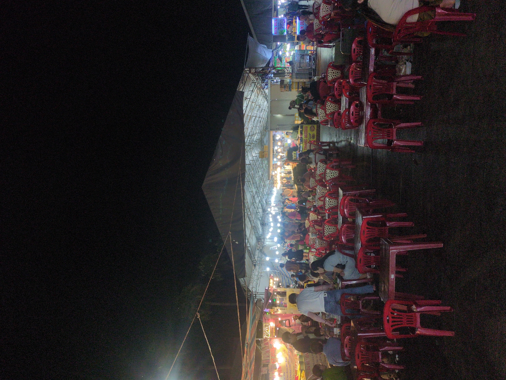The image depicts a bustling outdoor dining area, likely a street food market, in the city of Tuy Hòa, Vietnam, at night. Long rows of red plastic tables and chairs are occupied by people eating and socializing. Overhead, a makeshift roof structure with metal supports and fabric covering is illuminated by numerous strings of lights. Various food stalls and signs, some in Vietnamese, are visible. The overall atmosphere suggests a lively and informal dining experience.