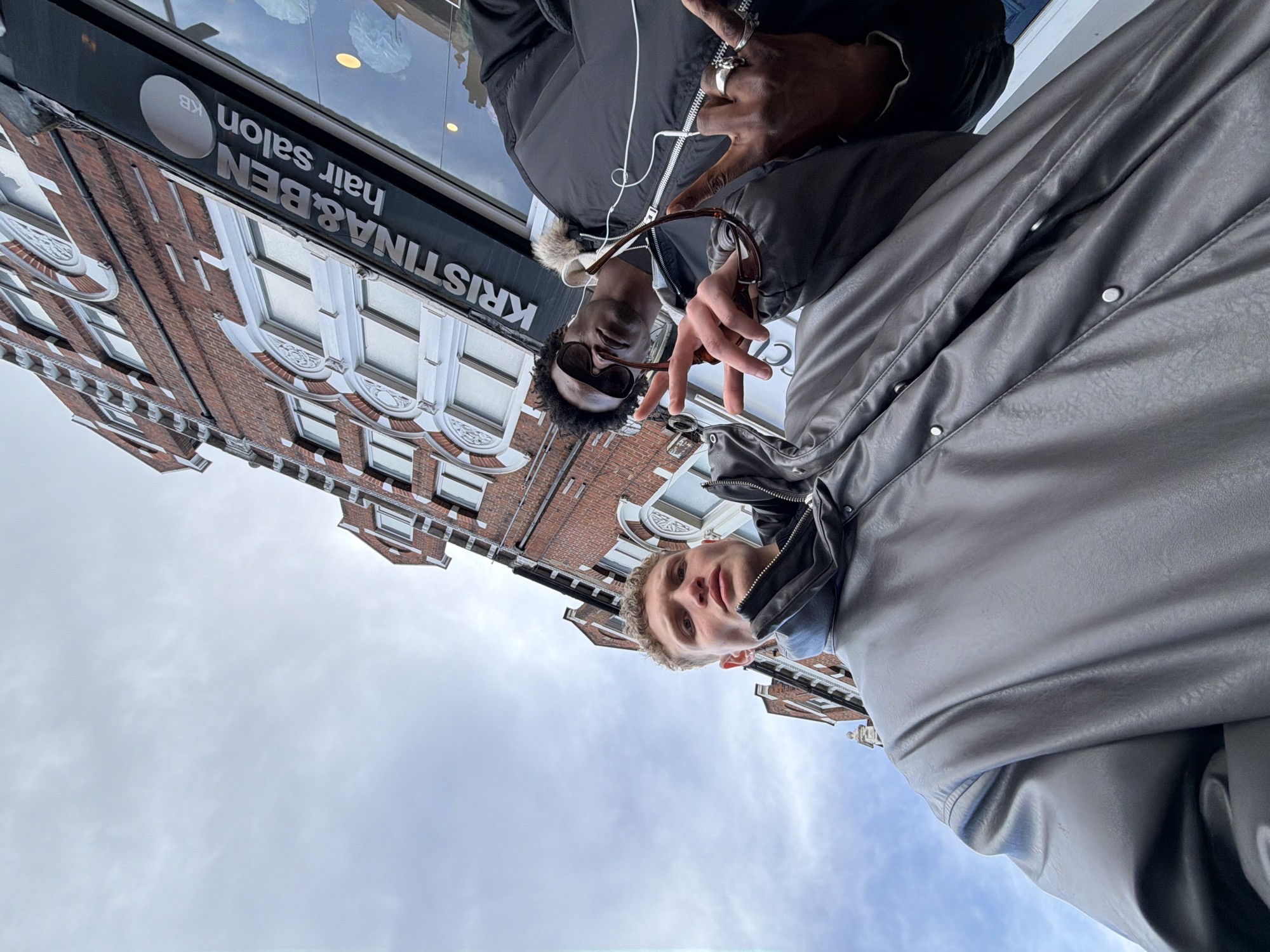 A low-angle, wide-perspective photograph depicts two young males posing in front of the Kristina & Ben Hair Salon, located at 122 Ladbroke Grove, London W11 1BP, United Kingdom, beneath an overcast sky.
The subject positioned on the left is a fair-skinned male with short, light-brown curly hair. He wears a dark grey, textured puffer-style jacket with a prominent high collar and looks directly at the camera with a neutral expression.
The subject on the right is a dark-skinned male wearing dark sunglasses and a black puffer jacket featuring a visible fur-trimmed hood. His left hand holds a pair of brown-framed sunglasses, partially obscuring his face. His right hand is raised, forming a 'peace' sign, and displays multiple silver rings on his thumb, index, ring, and pinky fingers. A white earbud wire is visible draped over his jacket.
The background consists of a multi-story red brick building characterized by white, ornate architectural details around its windows. A black, vertically oriented sign with the white text "KRISTINA & BEN hair salon" is affixed to the building's facade, adjacent to a reflective glass storefront.