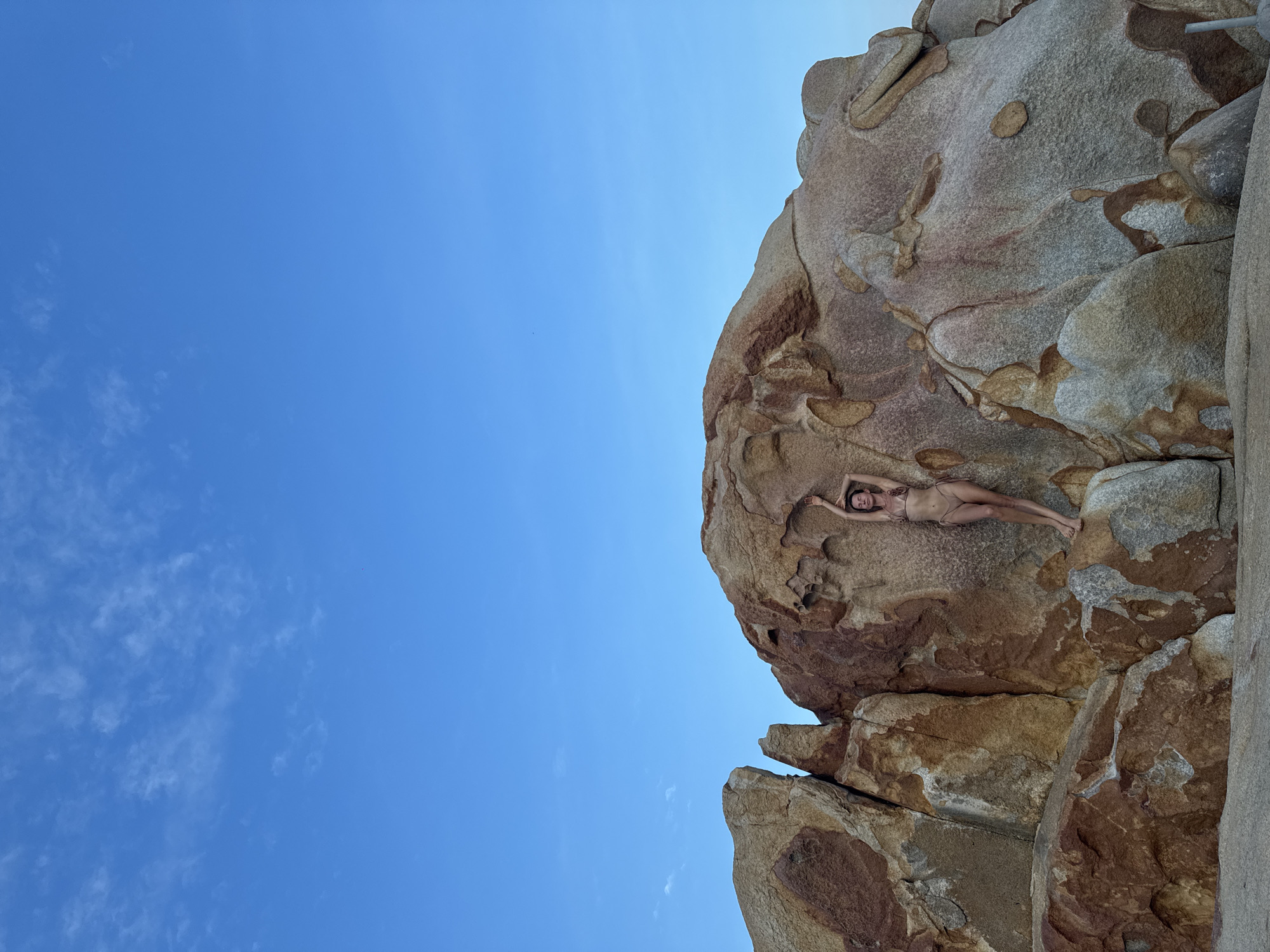 A woman in a bikini poses with her arms raised against a large, weathered rock formation under a clear blue sky. The rock is predominantly reddish-brown and gray, with visible textures and natural indentations. The woman is positioned on the right side of the frame, leaning against the rock. The foreground shows the sandy or rocky ground. The background consists of a vast expanse of blue sky with a few wispy clouds. The scene appears to be a coastal or natural landscape. This location is on the coast of Thuận Thành, Vietnam.