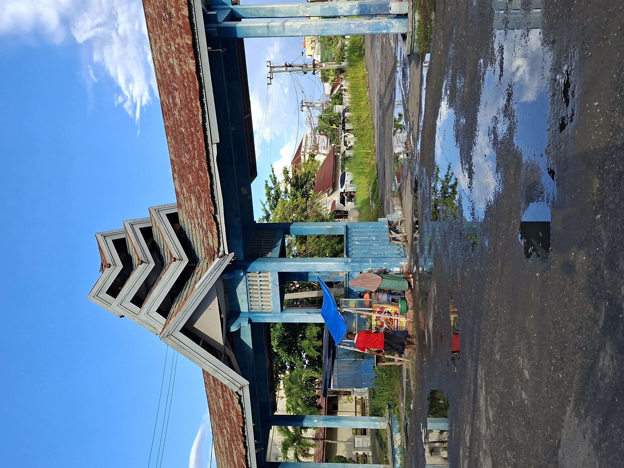The image captures a scene in Lhokseumawe, Indonesia. A man wearing a red shirt and a dark sarong-like garment stands next to a colorful vendor cart, possibly selling food or drinks. A blue flag or cloth hangs near the cart. The vendor and cart are situated near a blue-painted structure, which appears to be the entrance to a building or compound. The structure has a distinctive stepped gable design. The corrugated iron roof is rusted.
The ground is wet, with puddles reflecting the sky and surrounding buildings. In the background, buildings, power lines, and greenery can be seen. The sky is blue with some white clouds. The image orientation suggests it was taken from a slightly unusual angle.