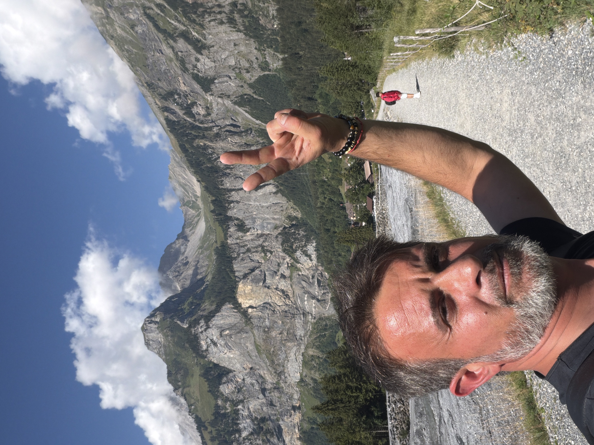 A man with graying hair and a beard takes a selfie in front of a mountainous landscape. He is wearing a black shirt and has his right arm raised, forming a peace sign with two fingers. On his wrist, he wears several bracelets. In the background, a person in a red jacket walks away down a gravel path. The path is lined with wooden posts and rope fencing. Beyond the path, there are evergreen trees and then a steep, rocky mountain face. The sky is blue with scattered white clouds. The scene appears to be in Kandersteg, Switzerland, a location known for its alpine scenery and hiking trails.