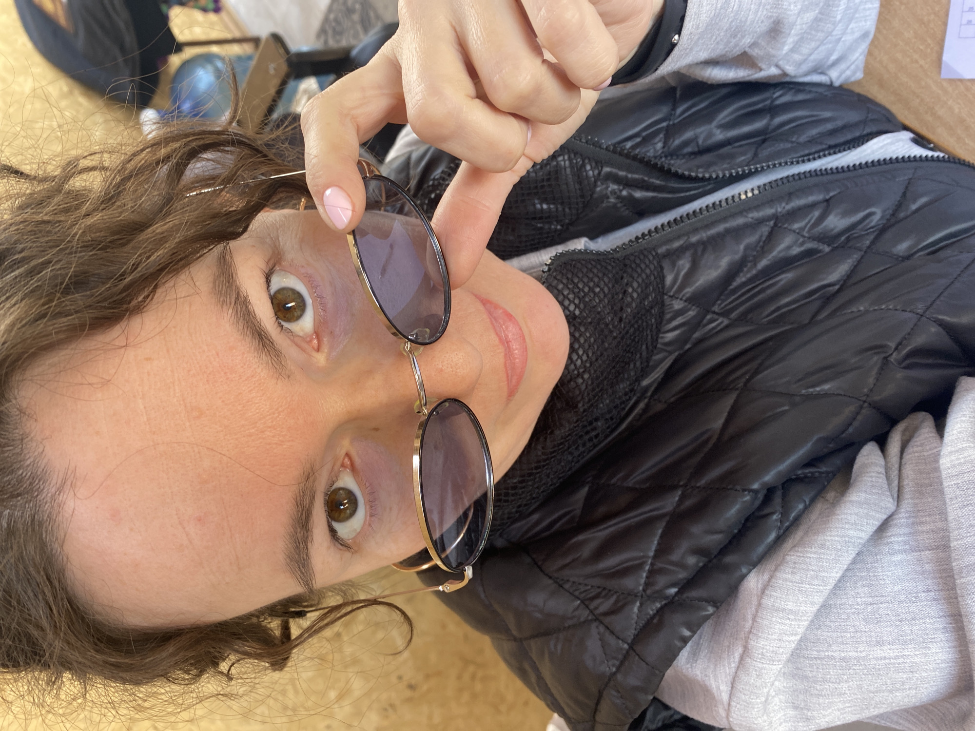 A woman with brown hair and hazel eyes holds a pair of sunglasses up to her face. She is wearing a black quilted jacket over a gray hooded sweatshirt. The background is blurred, but a globe is visible. The location is presumed to be Ol'khovskoye, Russia.