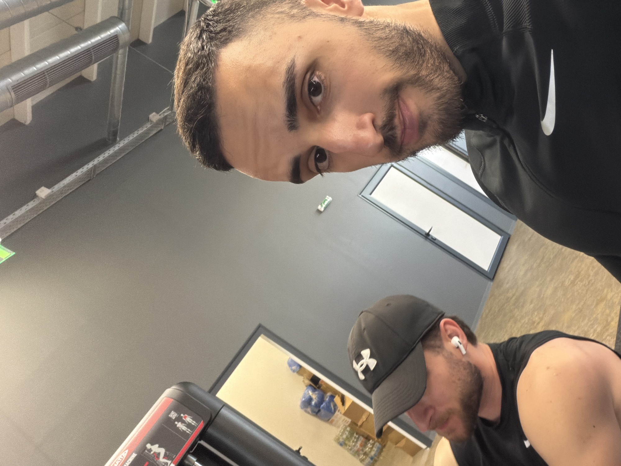The image appears to be taken indoors, possibly in a gym or fitness center in Plovdiv, Bulgaria. Two men are visible. The man closer to the camera has dark hair, a beard, and is wearing a black Nike jacket. He seems to be taking a selfie. The second man is wearing a black Under Armour baseball cap and a black sleeveless shirt. He has a beard and an earbud in his ear. A weight machine is visible in the lower left of the frame. Shelves with equipment are in the background. The environment appears to be a modern, well-lit facility.