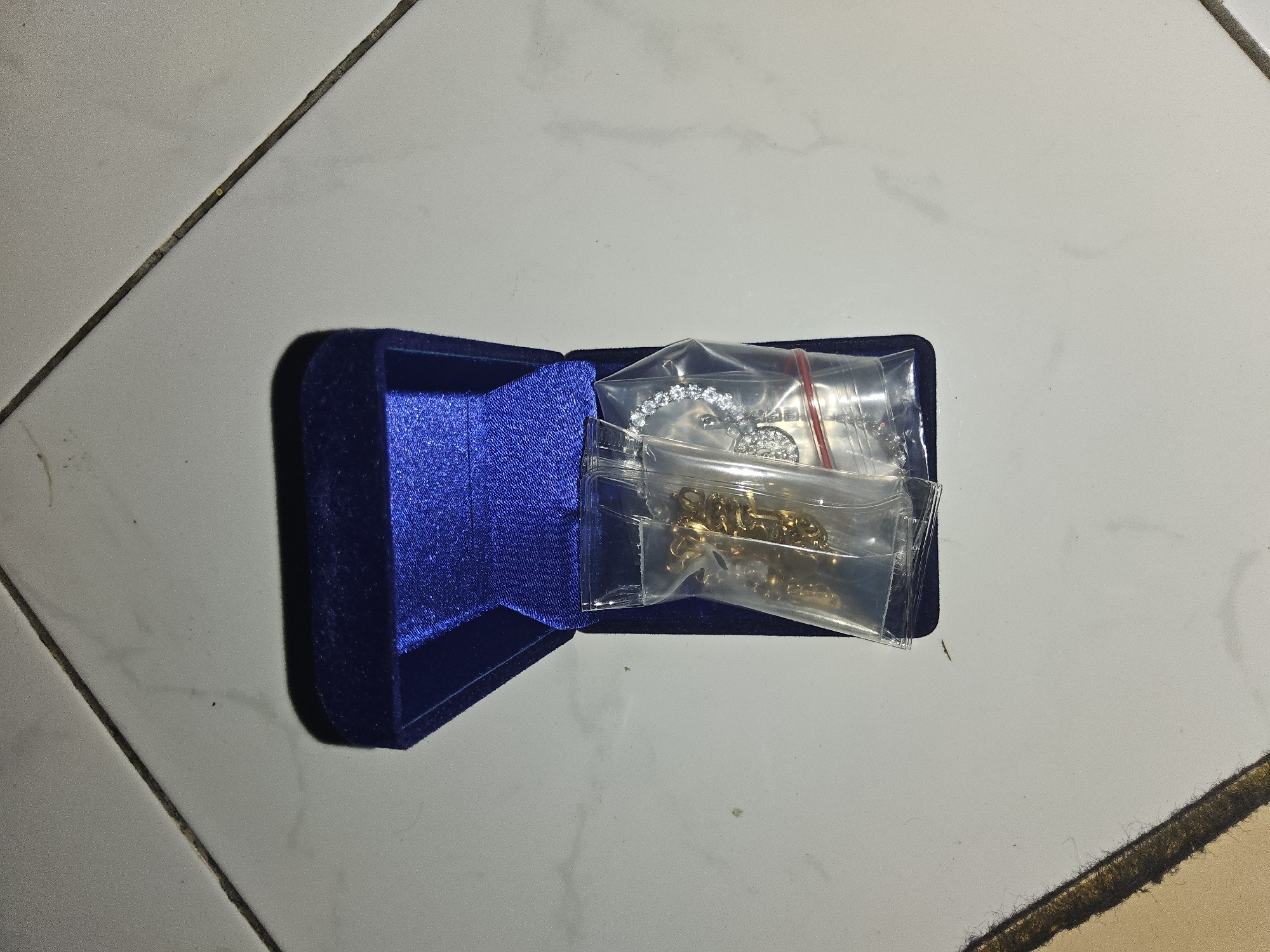 An open, dark blue velvet jewelry box sits on a tiled floor. Inside the box, two small plastic bags contain jewelry. One bag holds a gold-colored chain. The other bag contains a silver-colored bracelet with clear stones. The tiled floor appears to be white with dark grout lines. The image is well-lit, and the objects are in focus. The scene is indoors, likely in a residential setting. There are no people visible in the image. The overall setting is domestic and unremarkable.