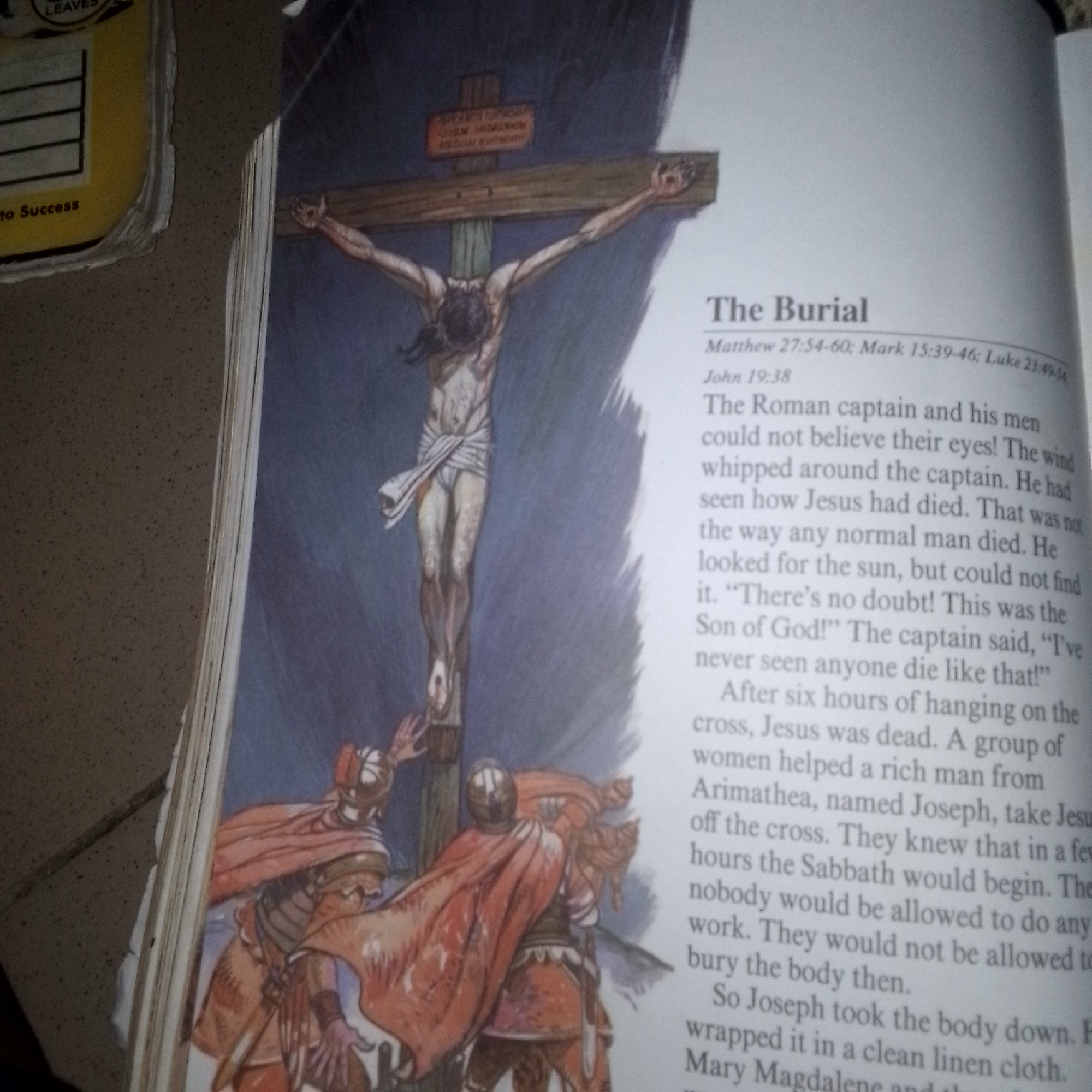 This image shows an illustration depicting the crucifixion of Jesus Christ. Jesus is shown on a wooden cross, with his arms outstretched and his body draped in a white cloth. The scene is dimly lit, suggesting it might be late in the day, and the overall mood is somber. Below the cross, several figures in red cloaks and helmets are gathered, appearing to be Roman soldiers or mourners.
To the right of the illustration, text from a book is visible. The heading "The Burial" is prominent, followed by biblical references: Matthew 27:54-60, Mark 15:39-46, Luke 23:49-54, and John 19:38. The text describes the events following Jesus' death, mentioning a Roman captain who witnessed his death and expressed disbelief, and the subsequent actions of Joseph of Arimathea and a group of women to take Jesus' body from the cross and prepare it for burial before the Sabbath.
On the left edge of the image, a portion of another object is visible, possibly a book or a packaging material, with faded text including "LEAVES" and "to Success". The image is a photograph of an open book, with the pages slightly curved.