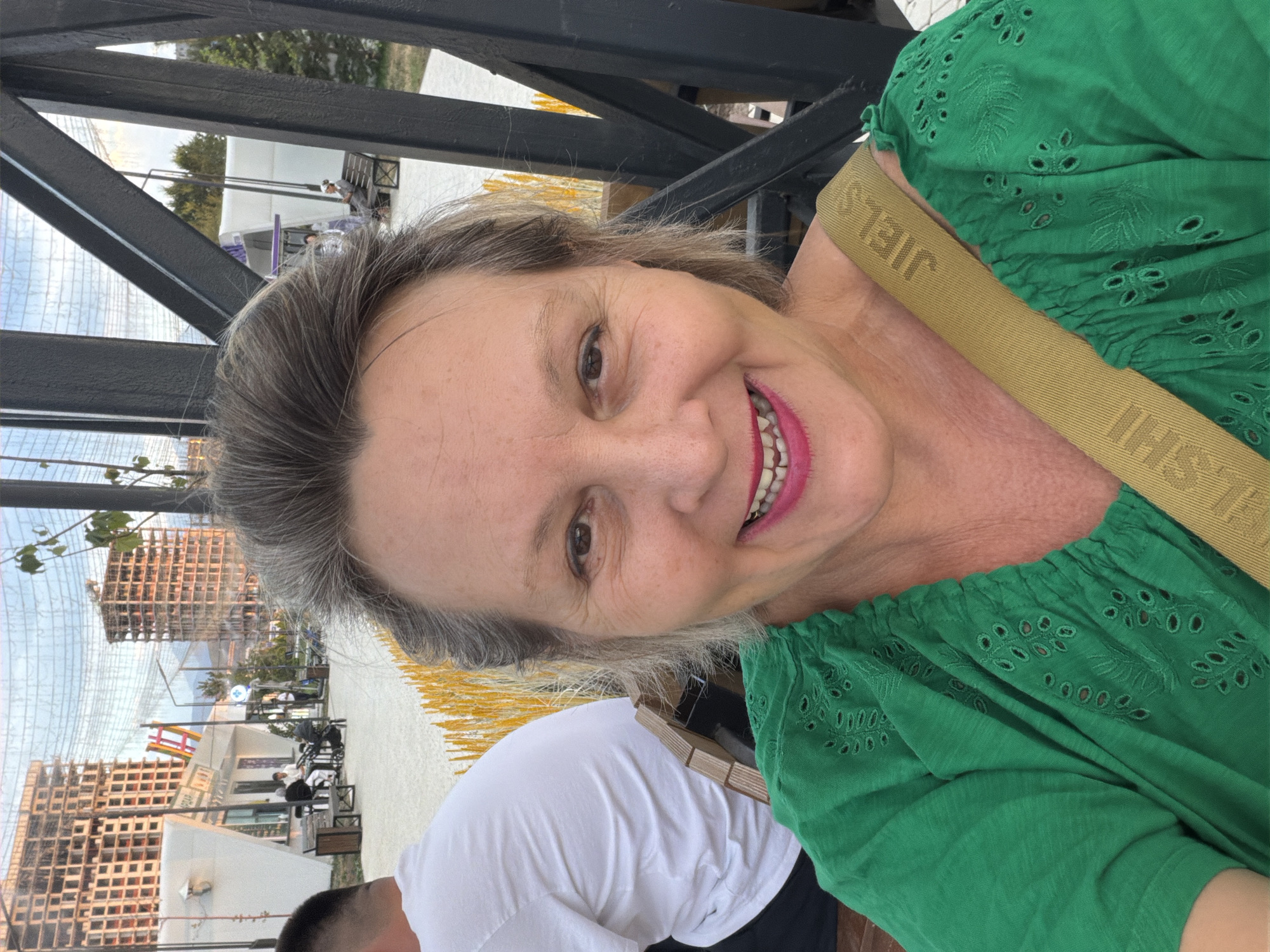 In an outdoor urban setting, a middle-aged woman with graying hair and a bright green top is taking a selfie. She is smiling broadly, showing her teeth. A tan strap with the word "JIELS" visible on it goes across her shoulder. Behind her, to her left, a man wearing a white t-shirt is visible from the back. In the background, there are unfinished buildings with exposed rebar, suggesting construction. There are also some white structures resembling kiosks or small buildings. The ground appears to be paved, and there are some yellow ornamental grasses. The overall scene suggests a public area under development.