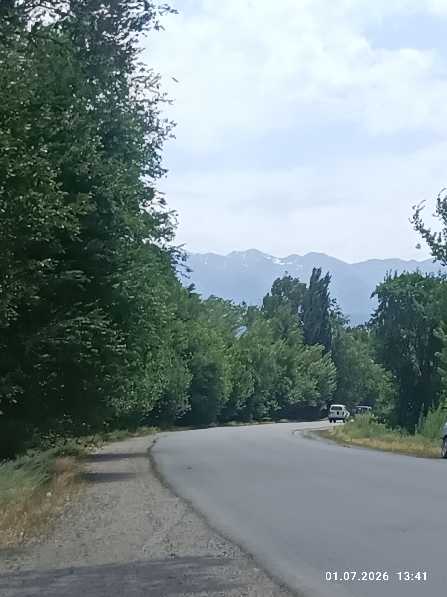 The image depicts a paved road curving to the left, bordered by a gravel shoulder on the left and dense green foliage and trees on both sides. In the distance, mountains with snow-capped peaks are visible under a partly cloudy sky. Several vehicles are present: a white SUV and another dark-colored vehicle are driving on the road in the distance. The timestamp "01.07.2026 13:41" is visible in the lower right corner of the image. The scene appears to be a rural or semi-rural area, possibly on the outskirts of Chüy, Kyrgyzstan, given the mountain range in the background.