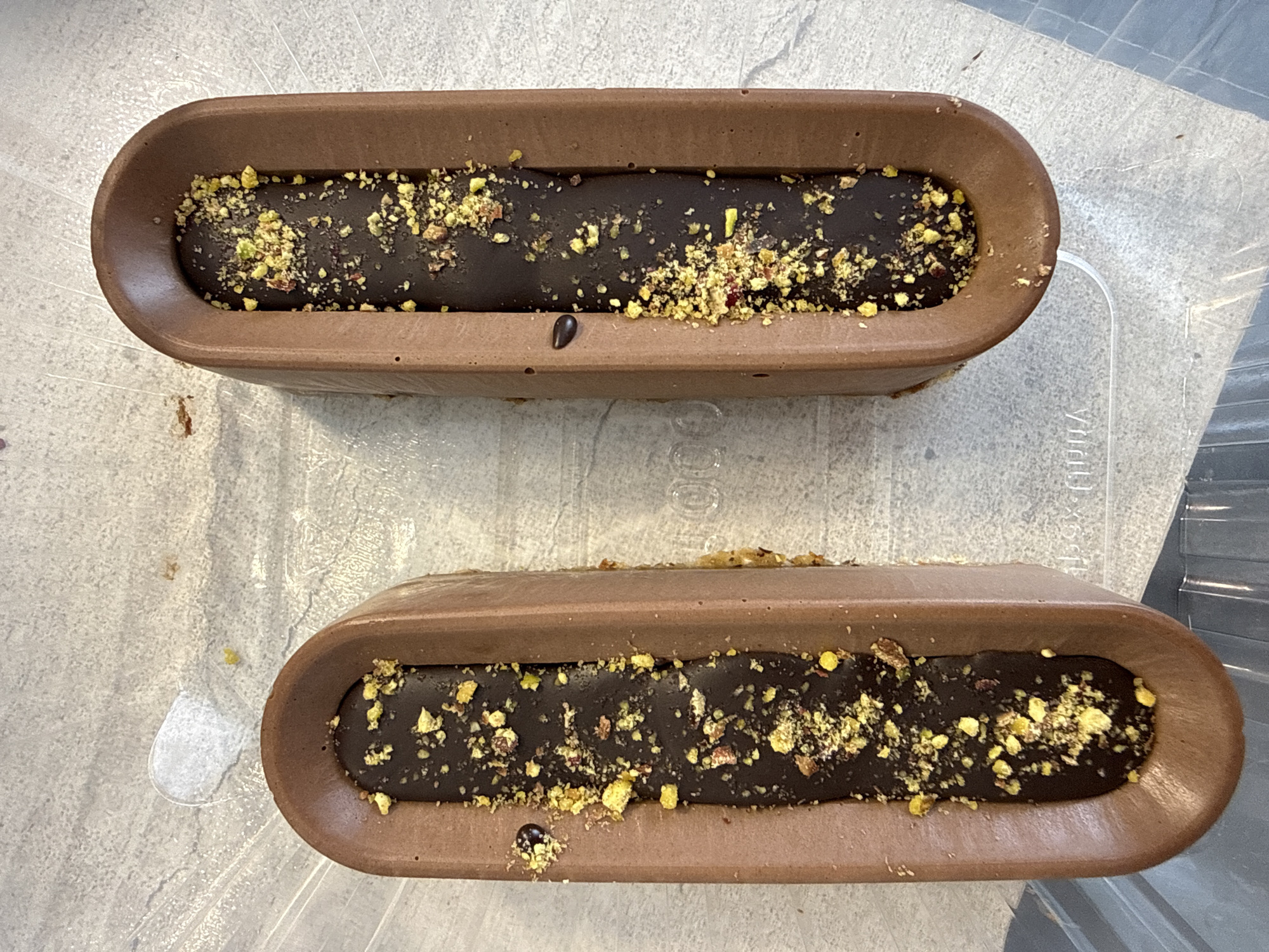 The image shows two oblong chocolate desserts. Each dessert has a molded, hollowed-out chocolate outer shell, filled with a dark, shiny chocolate filling and sprinkled with chopped pistachios. The desserts are placed side by side on a textured, possibly disposable, clear plastic surface. No people are visible in the image. The location in the city of Izhevsk, Russia is not possible to determine from the image provided.