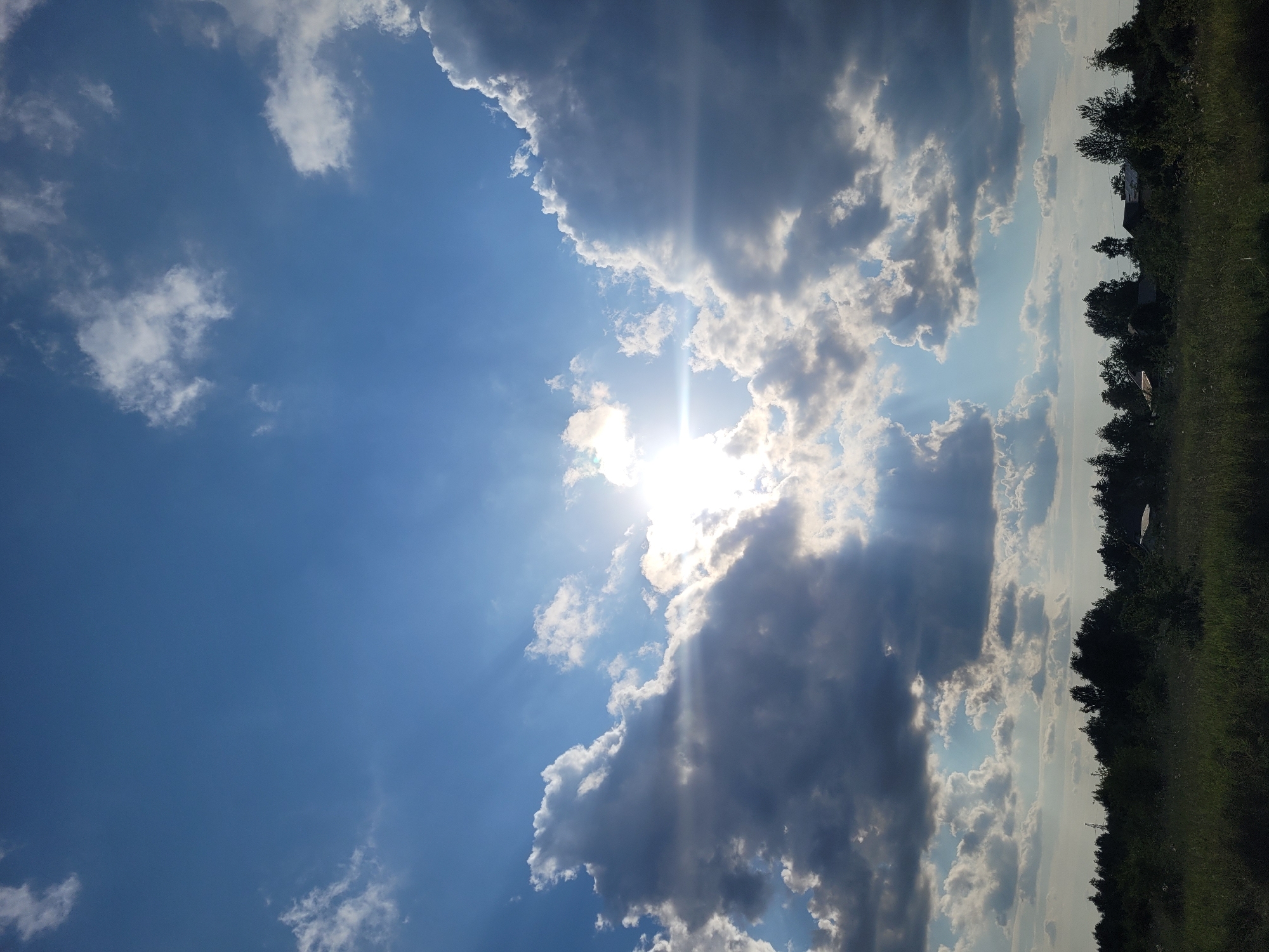 The image shows a landscape with a cloudy sky and a line of trees in the distance. The sun is visible through the clouds, casting rays of light. The foreground consists of a grassy field. There are no people or specific landmarks identifiable that would indicate the location as Jūrmala, Latvia.