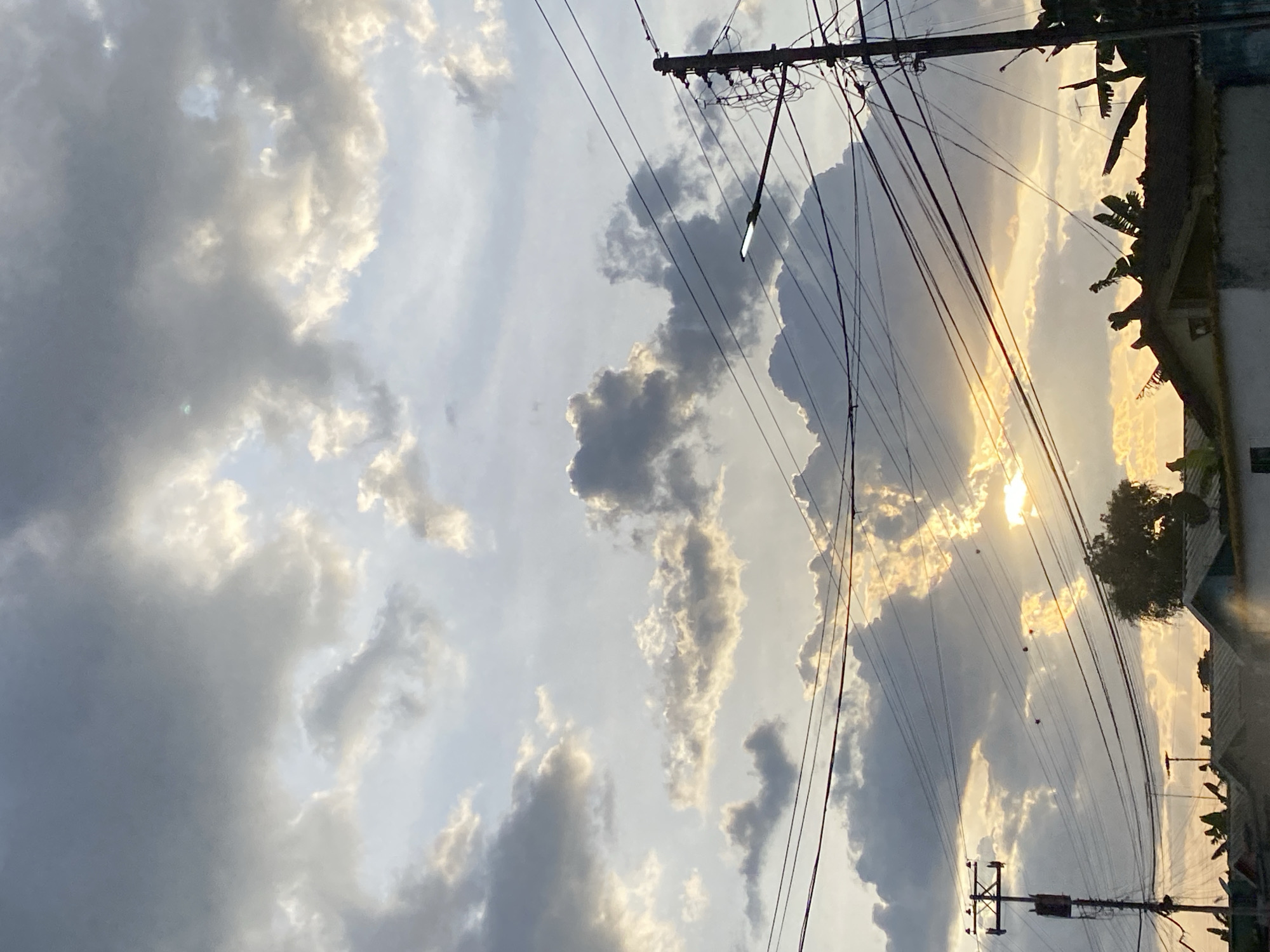 The image captures a sky with heavy cloud cover and visible sunlight. Electrical poles and wires are prominent in the foreground. Nearby rooftops and vegetation suggest a residential area. The location is potentially Tinaquillo, Venezuela, based on the image title.
